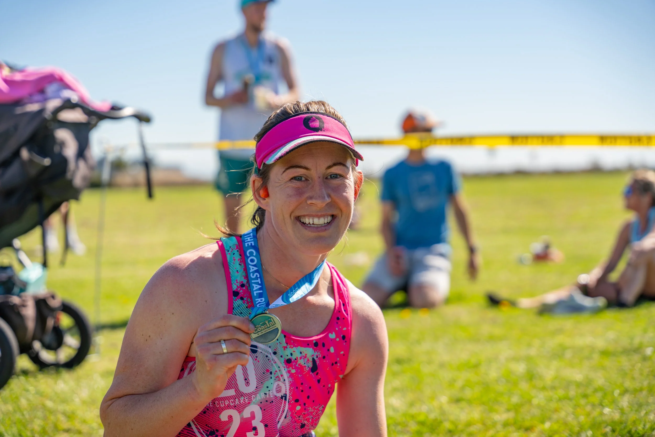 A smiling woman wearing a pink running outfit and a pink cap, holding a medal, after completing a race on a grassy field with spectators in the background.