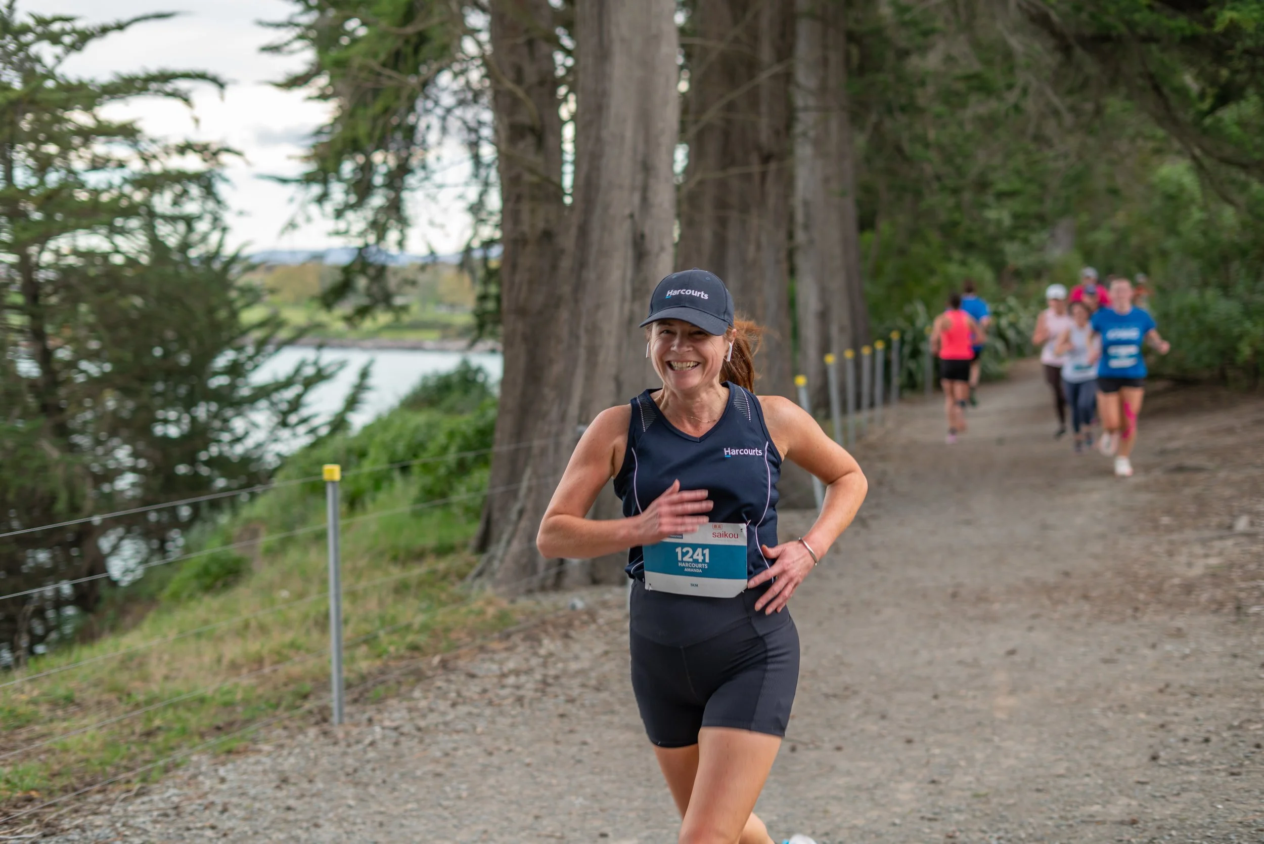 A woman running on a trail during a race, wearing a black cap and sleeveless black shirt with a bib number 1241, surrounded by trees and other runners in the background.