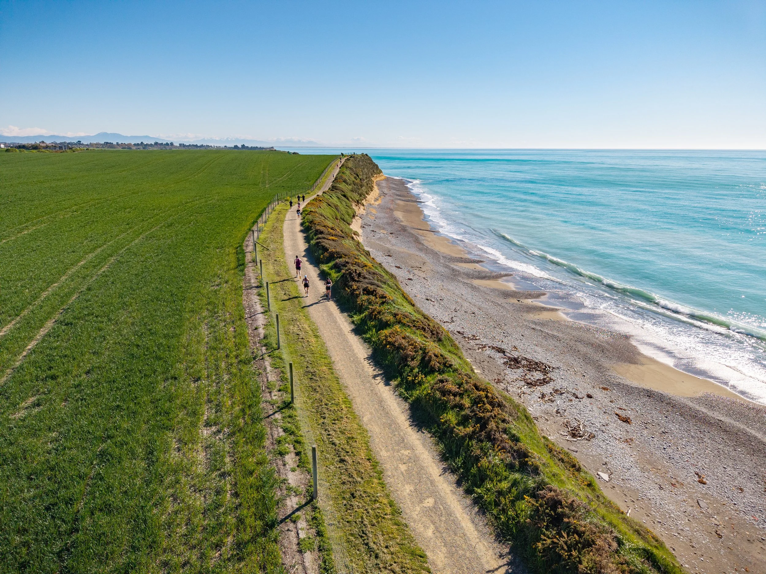 A coastal trail beside a beach with green fields on one side and the ocean on the other, with people walking along the path.
