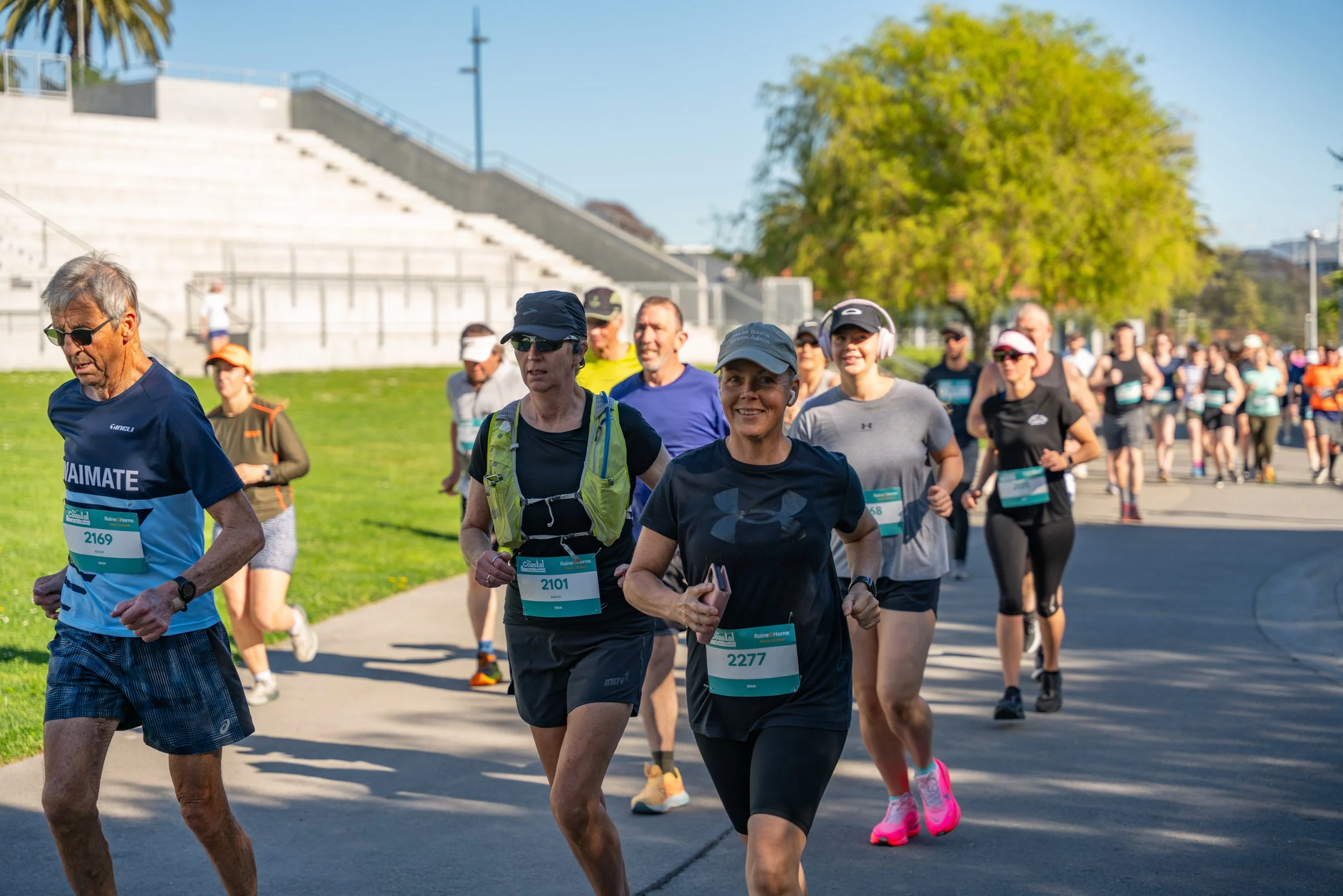 Group of people participating in a daytime outdoor running event, wearing athletic clothing, some with race bibs, on a paved pathway with grassy areas and trees, with bleachers and a blue sky in the background.