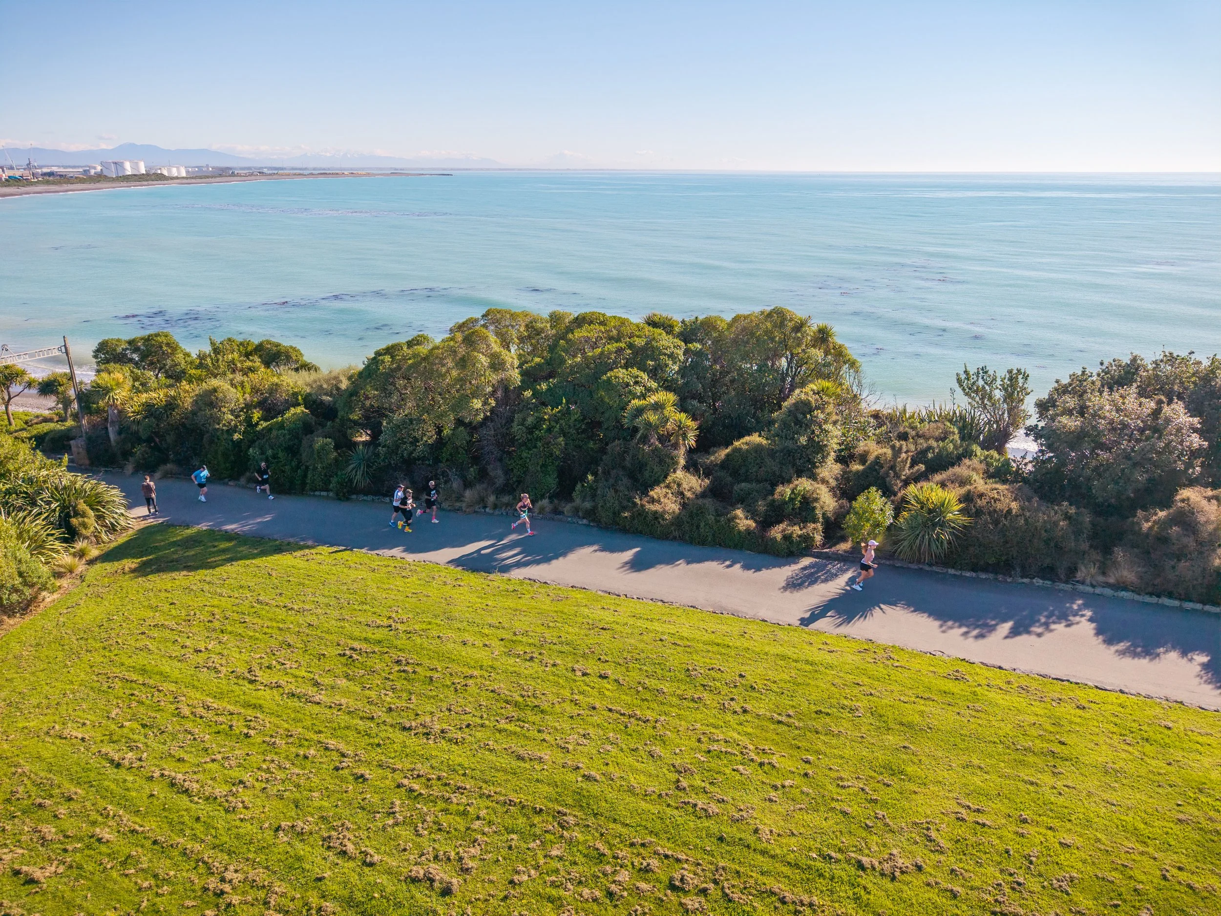 A group of people jogging along a coastal pathway near lush green trees with a view of the ocean and distant mountains.
