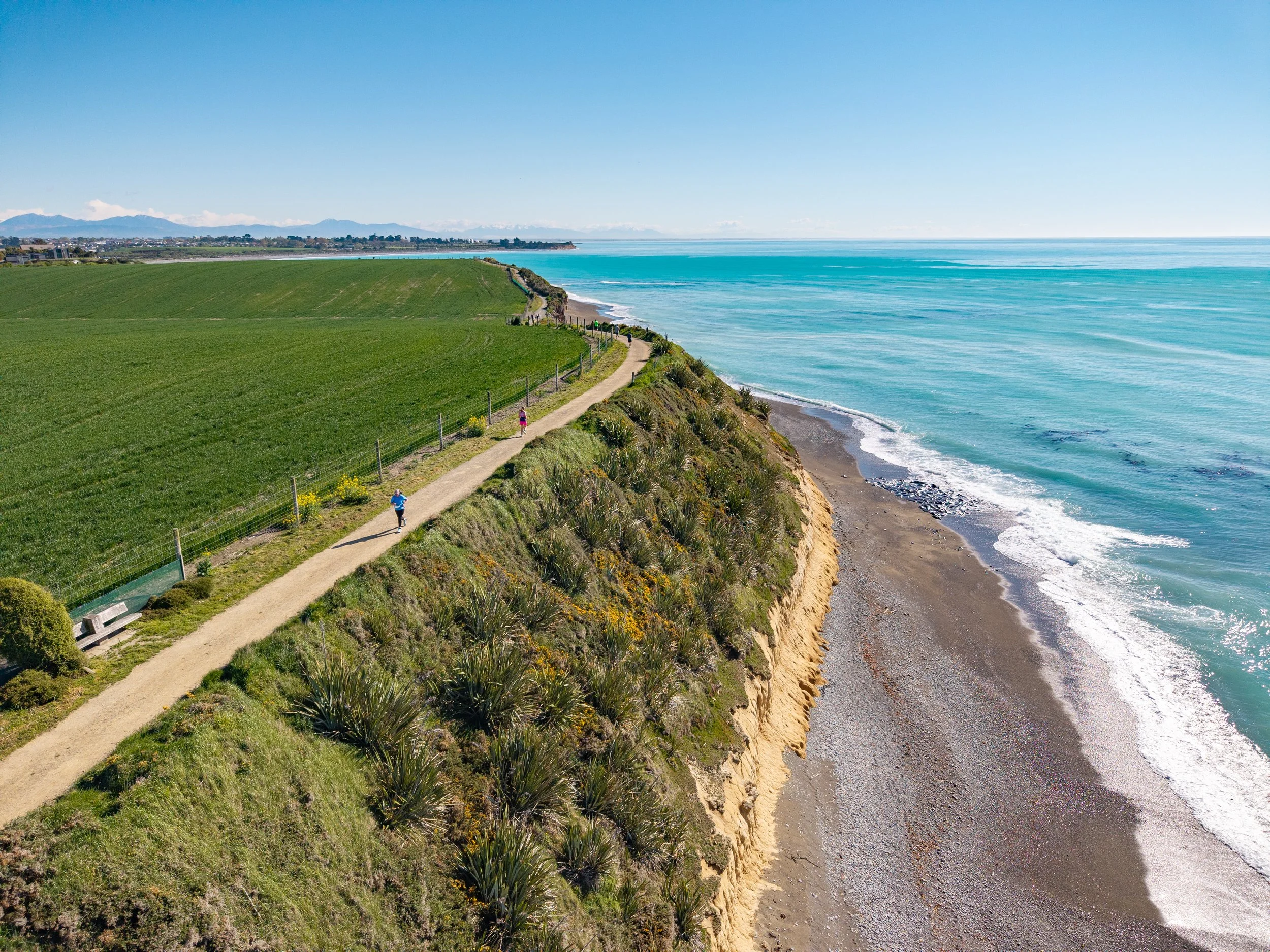 Aerial view of a coastal hiking trail along a green hillside near the ocean, with two people walking and a bench nearby, on a sunny day.