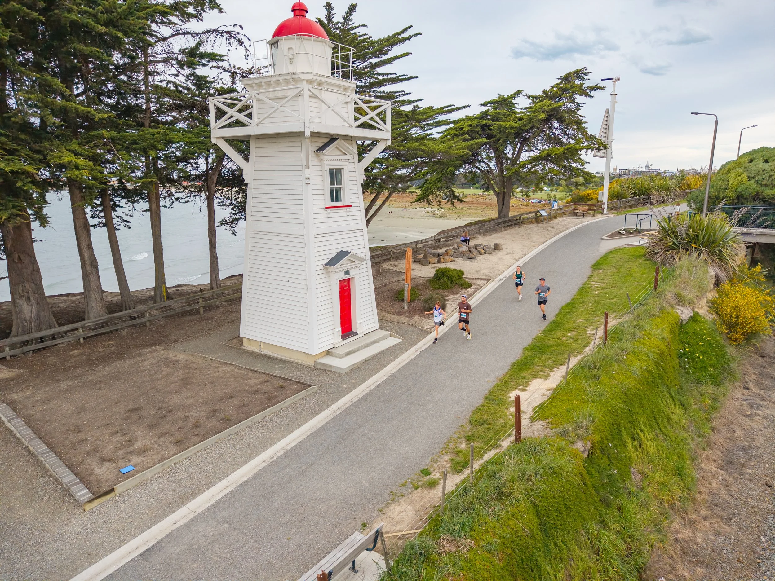A white lighthouse with a red roof next to a paved walking path, with five runners passing by. Behind the lighthouse are tall trees and the ocean.