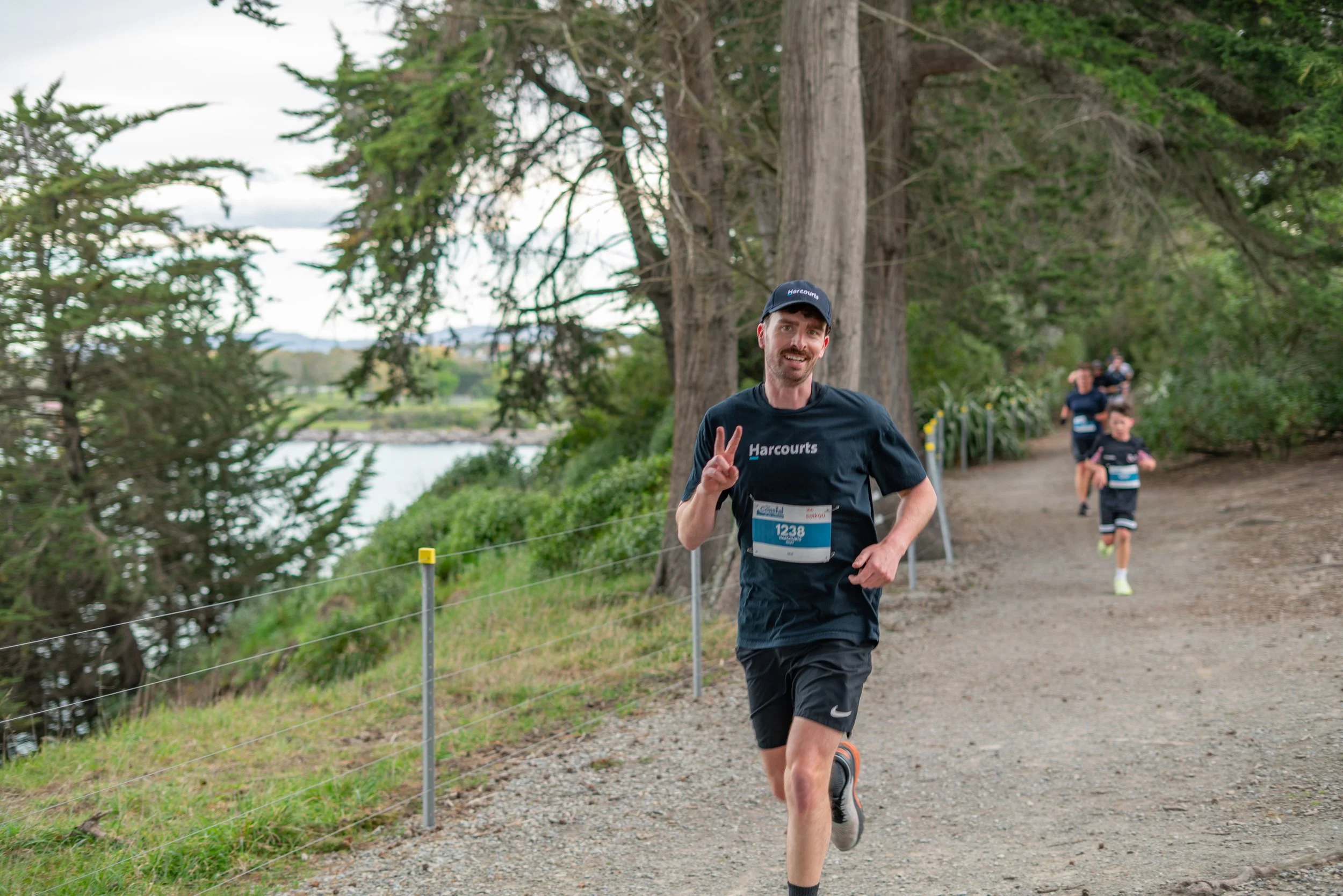 A man in black running clothes and a black cap making a peace sign, participating in a race along a wooded trail with other runners behind him.