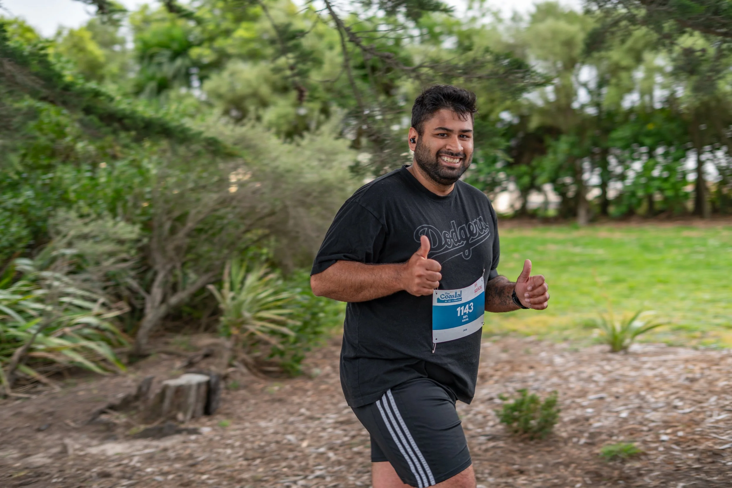 A man running outdoors, smiling, wearing a black t-shirt with "Dodgers" written on it, black shorts with white stripes, a race bib numbered 1143, and giving a thumbs-up gesture.