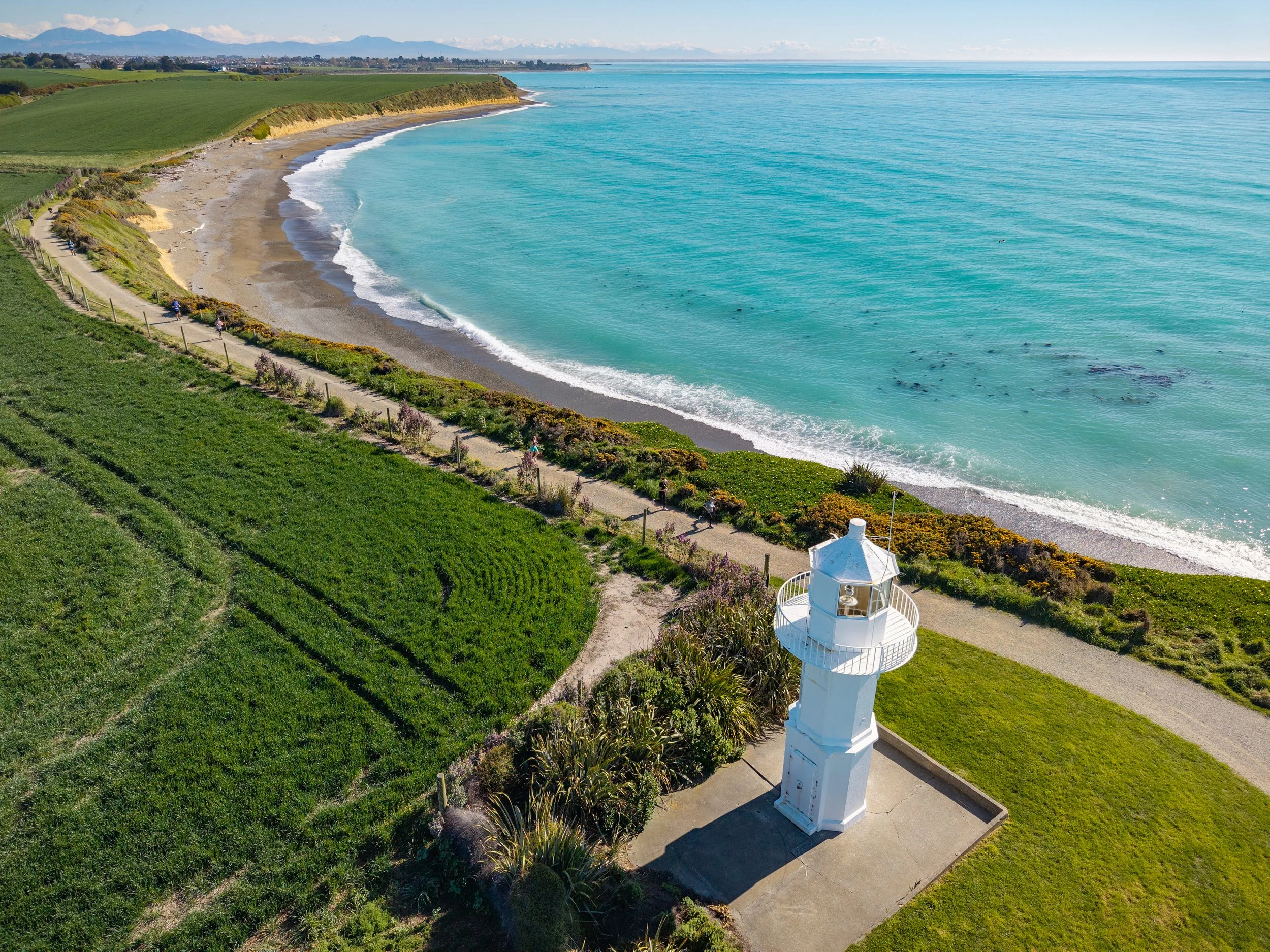 An aerial view of a coastal scene with a white lighthouse near the green grassy area and a pathway, a beach with waves, and the turquoise ocean stretching to the horizon.