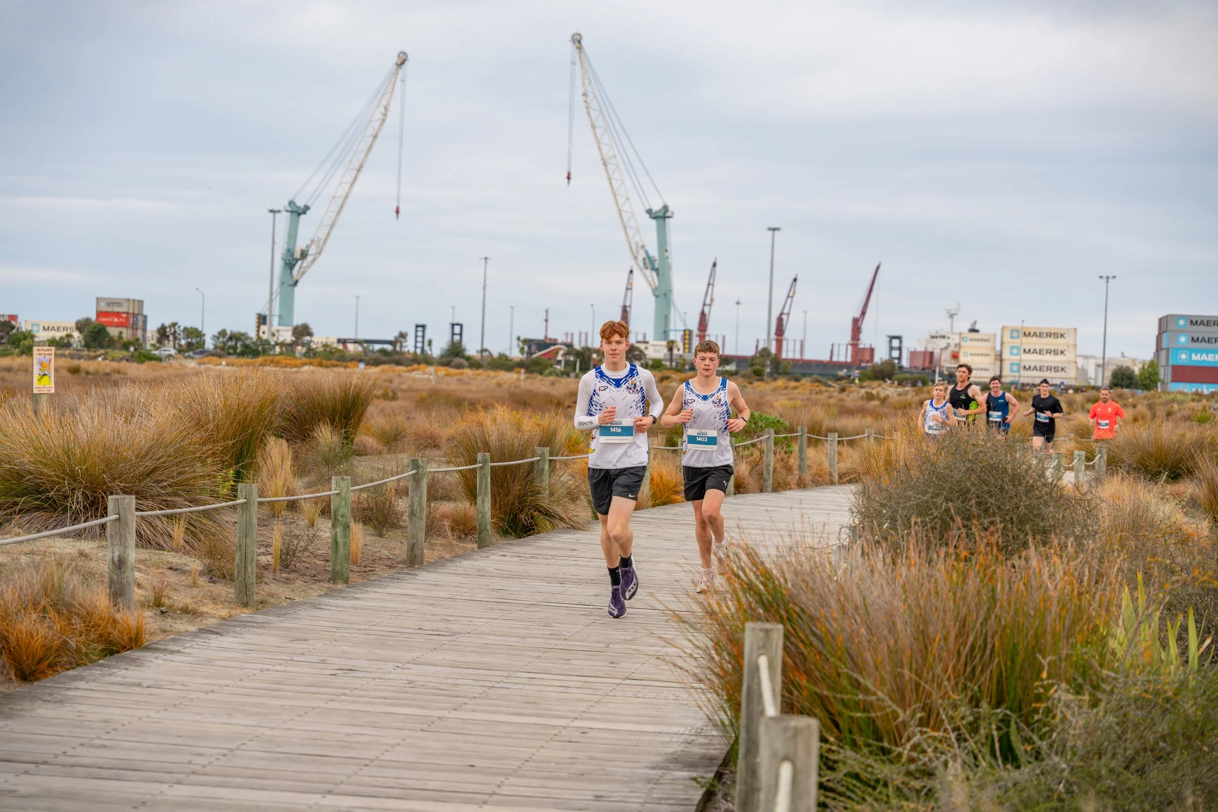 Two young men wearing athletic running clothes, participating in a race on a wooden pathway through a natural landscape with dry grass and bushes. A group of runners follows behind them. Industrial cranes and shipping containers are visible in the ba