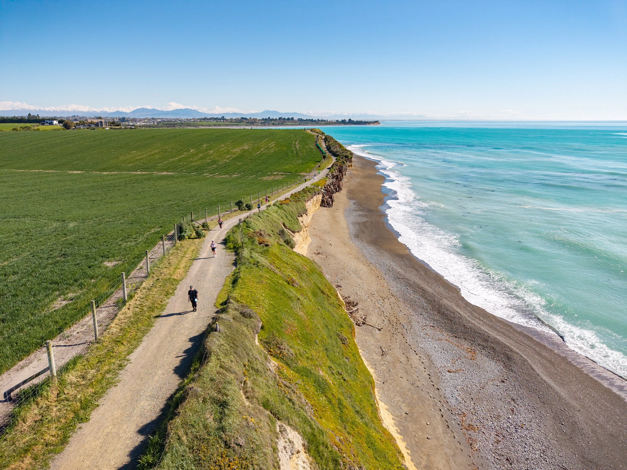 A coastal trail along a green cliff with people walking and biking, overlooking a beach and the ocean on a clear day.