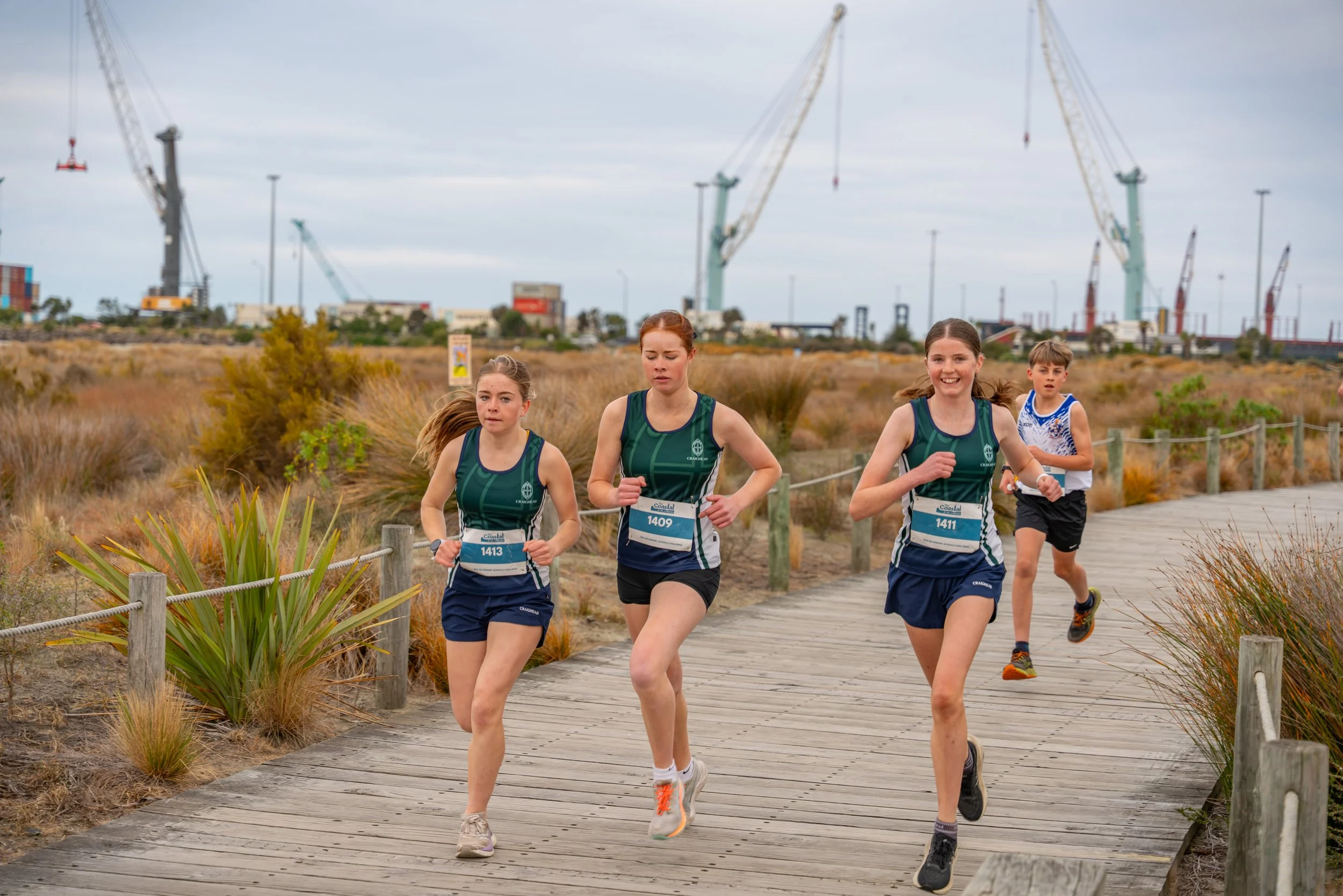 Four children running on a wooden trail in an outdoor area with plants and cranes in the background.