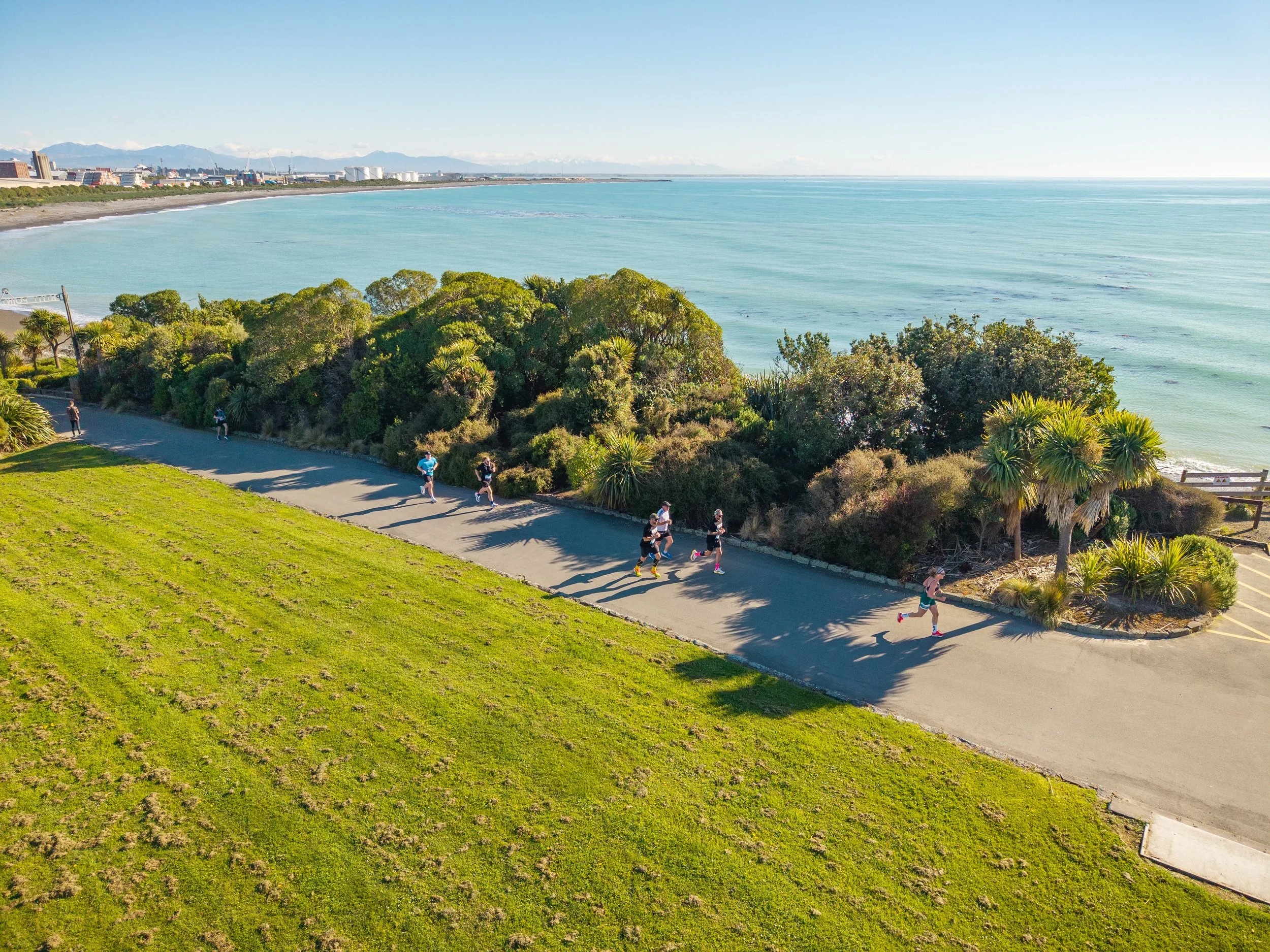 People jogging on a path along a green park with bushes and trees beside a beach and ocean, with a city skyline and snow-capped mountains in the background.