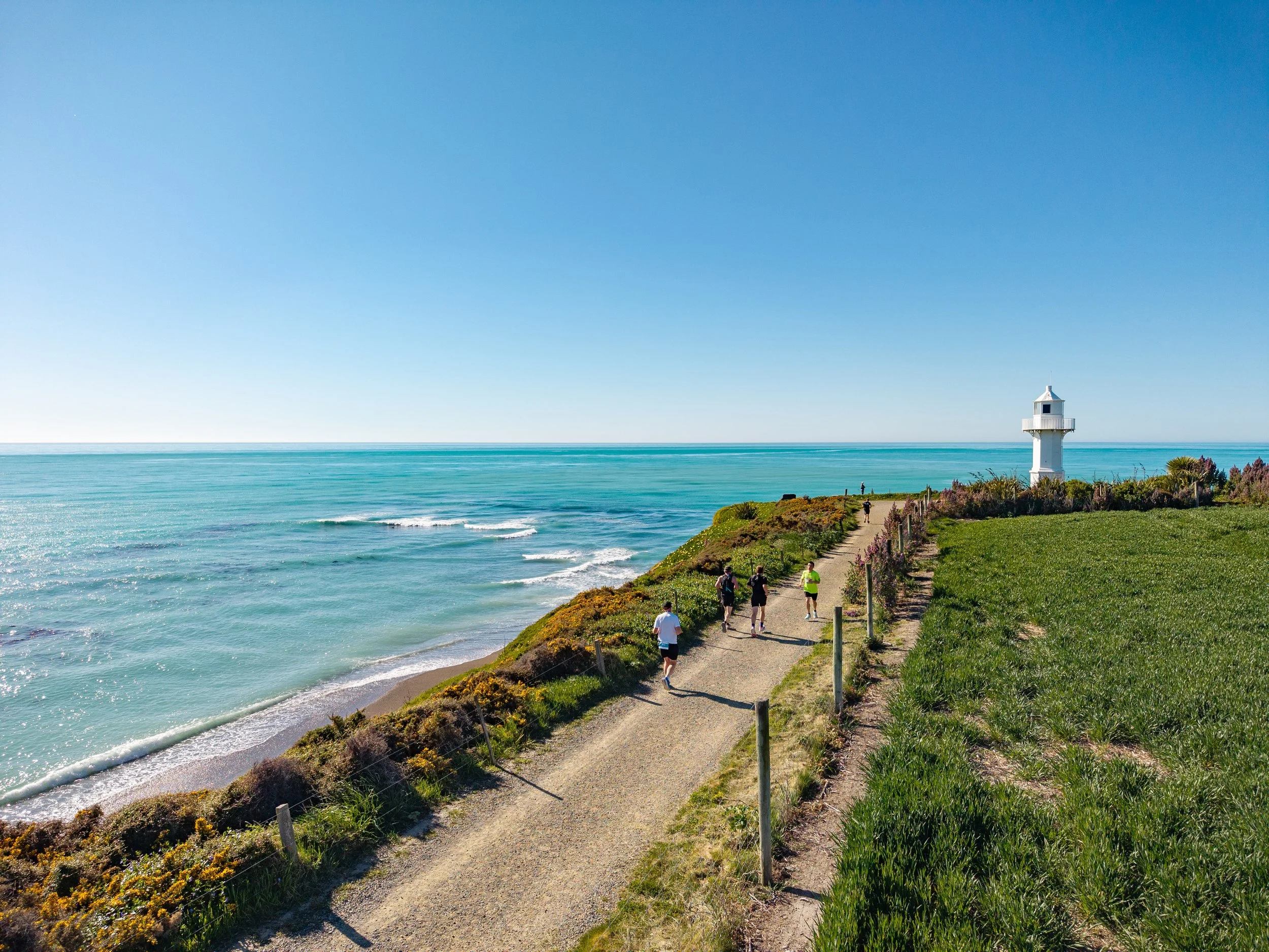 A coastal scene with a gravel path leading to a white lighthouse on a grassy hill, with six people walking towards the lighthouse, and the ocean and blue sky in the background.