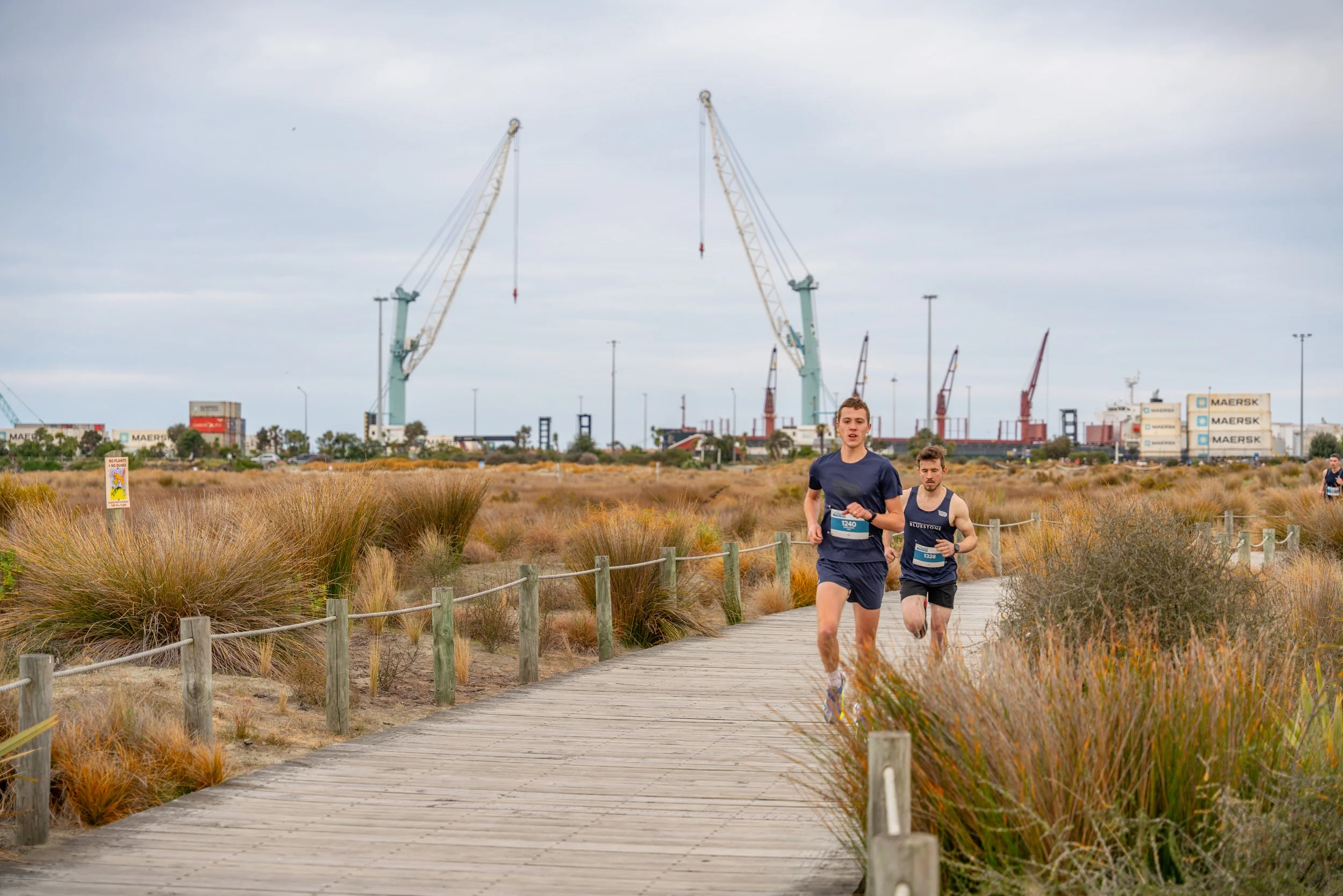 Two male runners participating in a race on a wooden pathway through a grassy area, with industrial cranes and shipping containers in the background under an overcast sky.