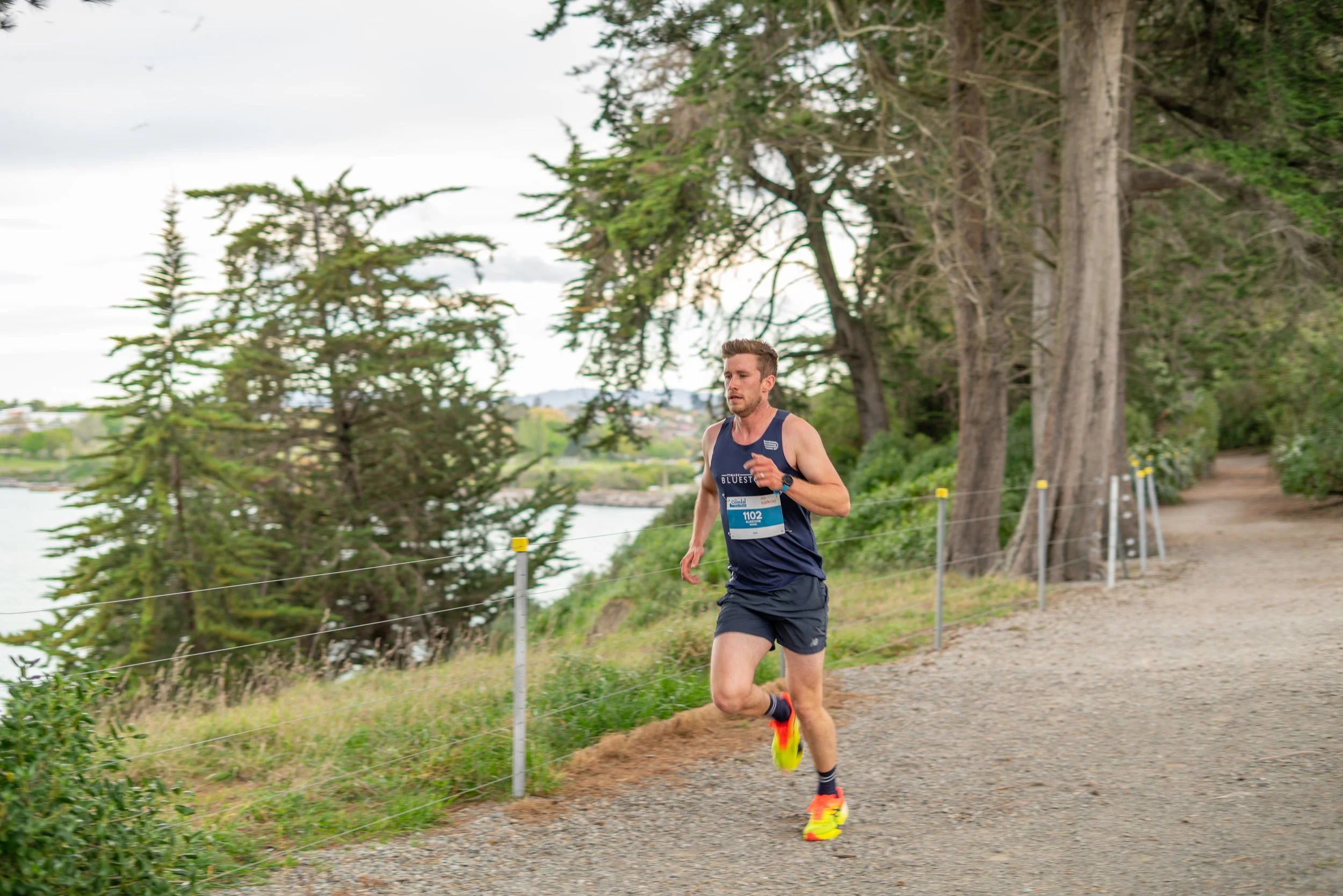 A man running on a trail by a body of water surrounded by trees during daytime.