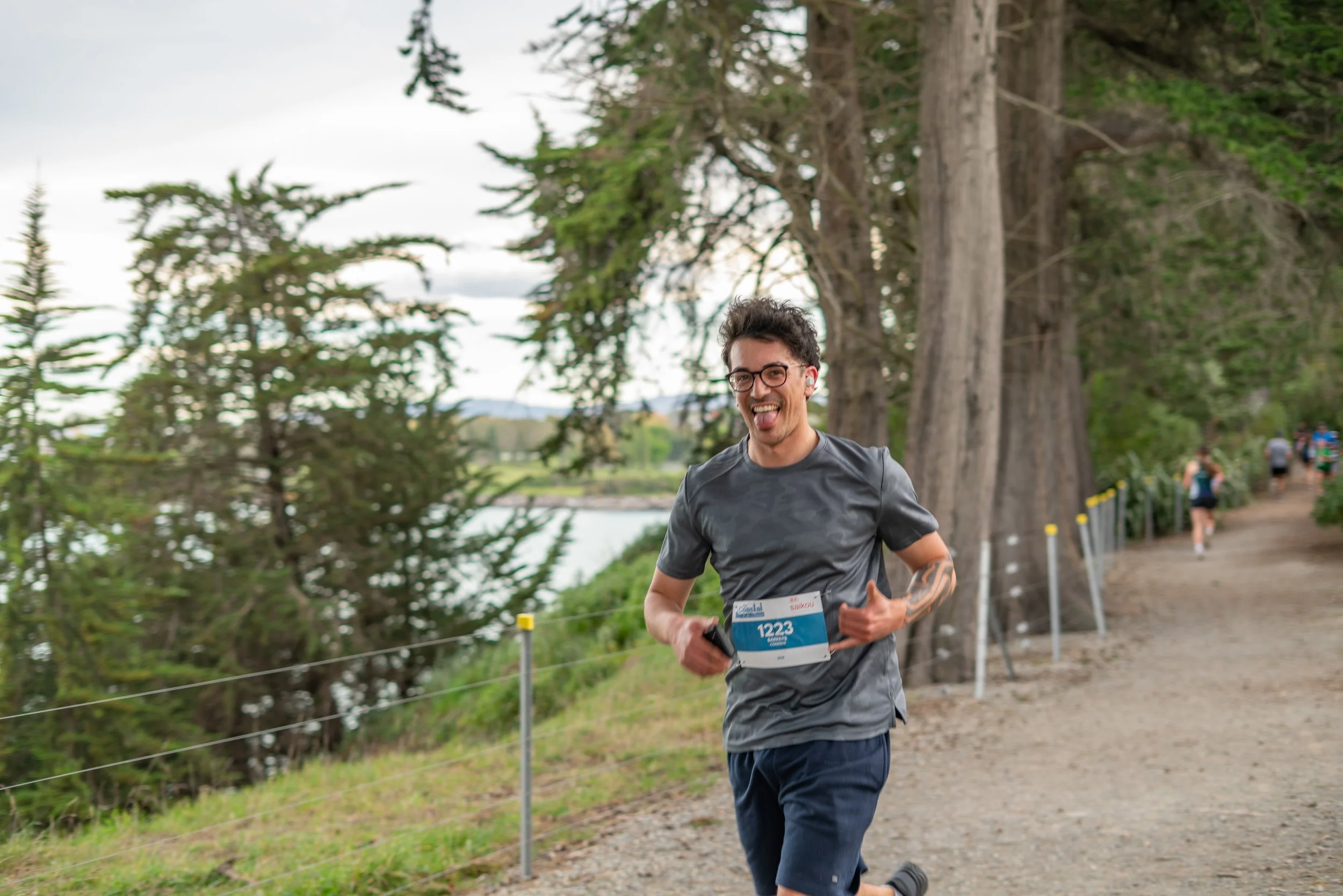 A man running outdoors on a trail with trees in the background, wearing a race bib numbered 1223, smiling and sticking out his tongue.