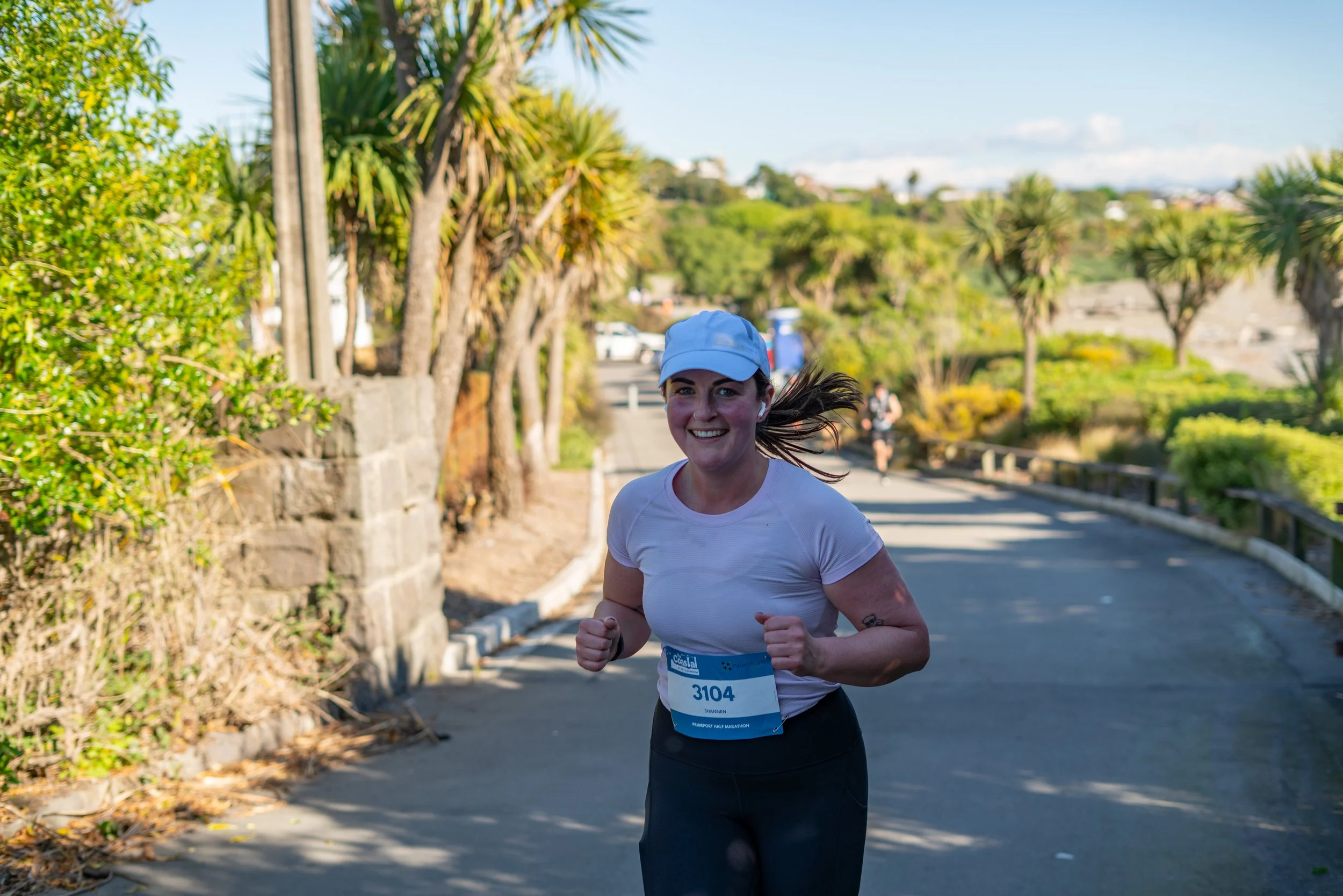 A young woman running outdoors on a sunny day, wearing a white cap, white shirt, and black leggings, with a race bib number 3104, smiling as she runs along a path with trees and greenery.