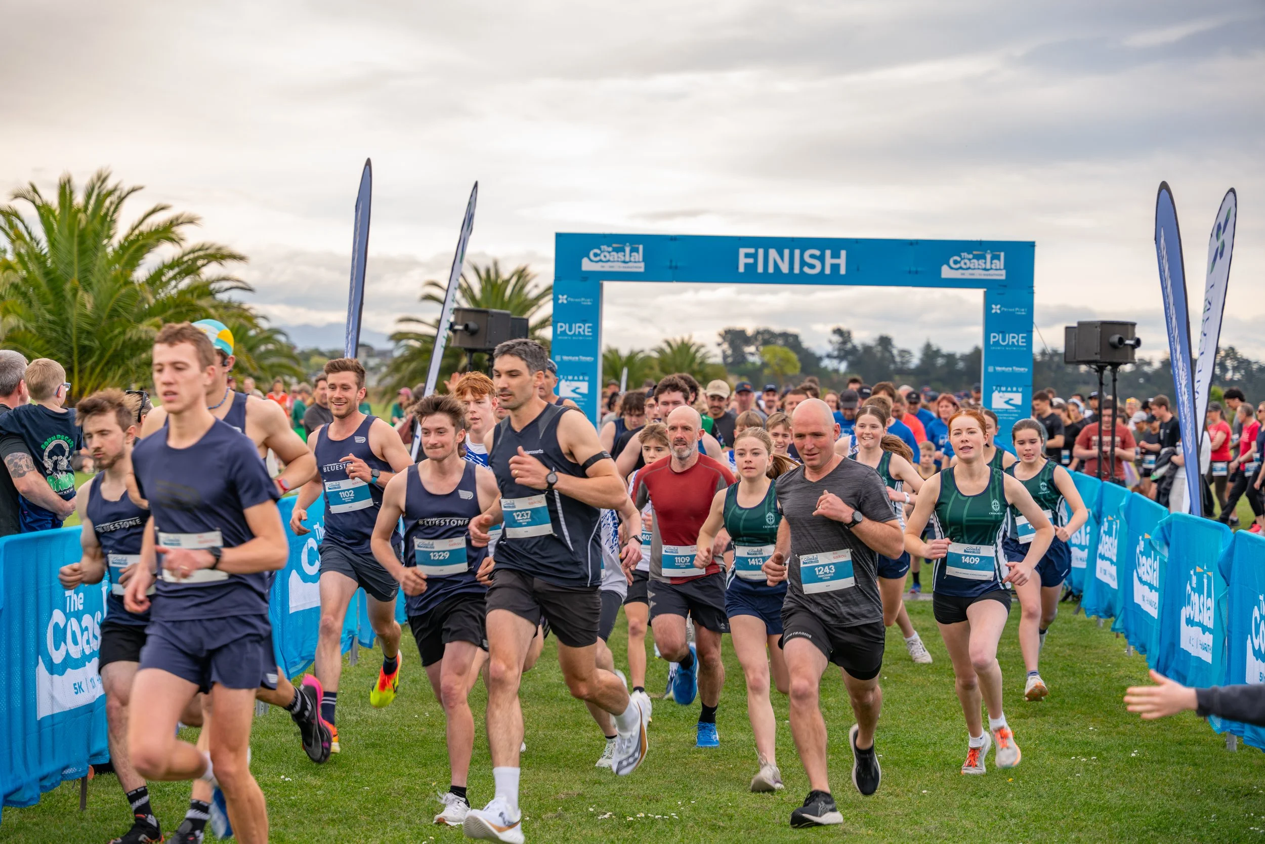 Group of runners crossing the finish line at a race event, with a blue finish banner overhead and a crowd in the background.