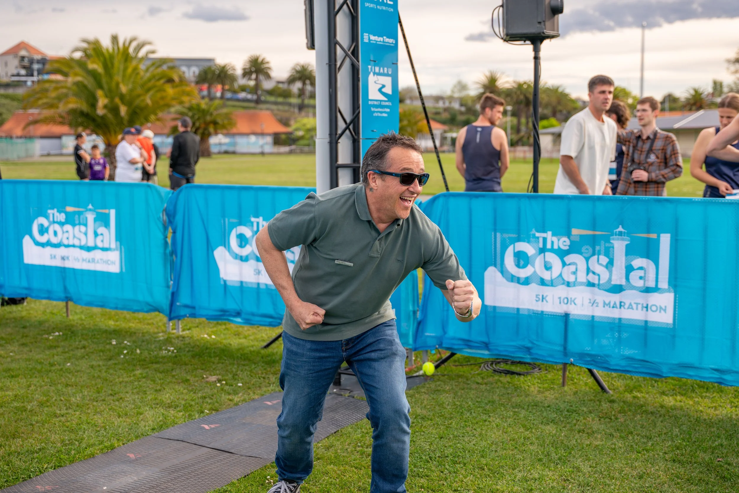 A man wearing sunglasses and a green polo shirt is celebrating at the finish line of a marathon, with a smile and fist pump, near blue barriers with 'The Coastal' marathon signage.