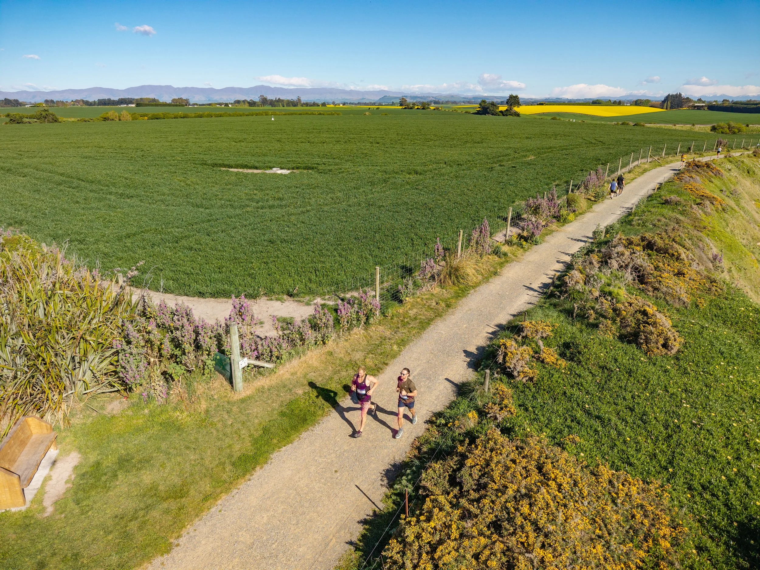 Two women jogging on a winding dirt trail alongside lush green fields and vibrant flowering bushes in a rural landscape with mountains in the background.