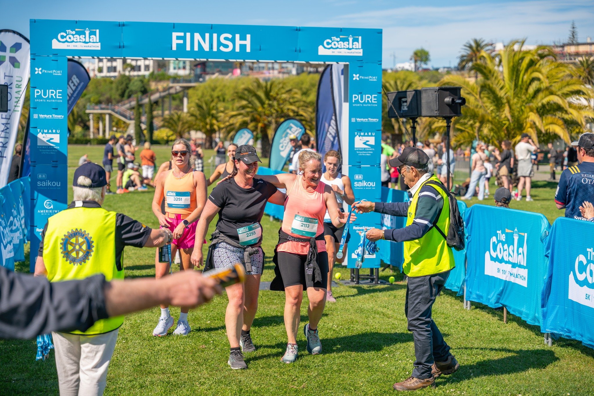 Runners crossing the finish line at a marathon event, with volunteers handing out medals and spectators in the background.