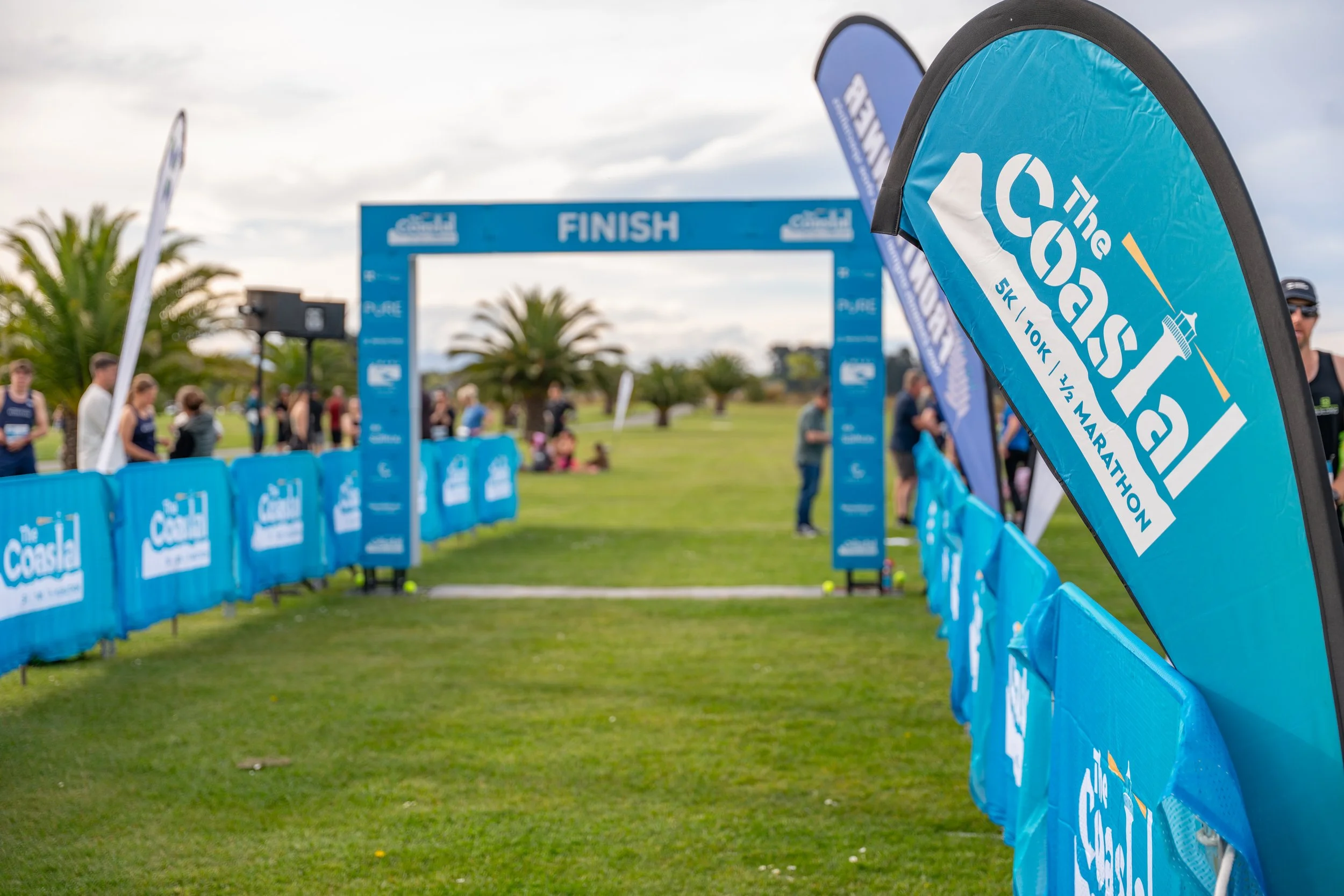 Finish line arch at a coastal marathon event with flags and banners, participants and spectators in the background on a cloudy day.