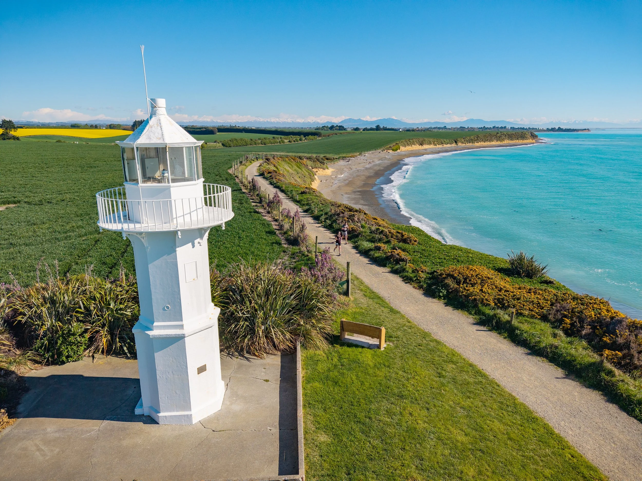 A white lighthouse stands at the edge of a grassy area overlooking the ocean, with a pathway running along the coast and people walking nearby. The scene features a sandy beach, green fields, and distant mountains in the background under a clear blue