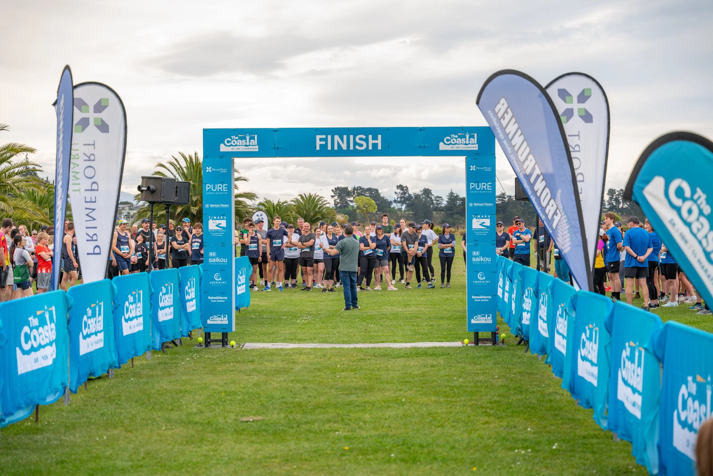 Participants gather at the starting line of a race outdoors, with a large blue finish gate and banners surrounding the area on a grassy field, under a cloudy sky.