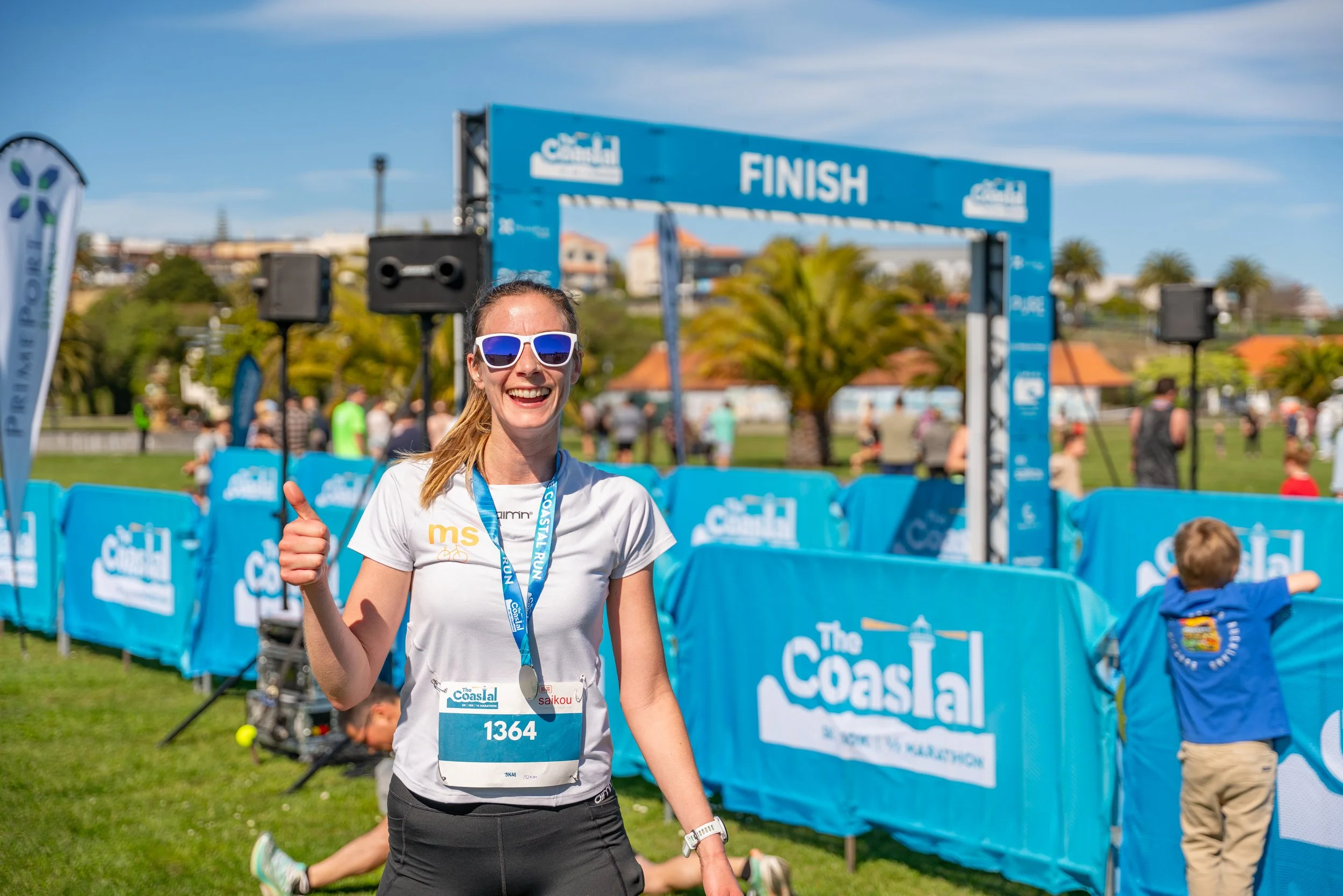 A woman smiling and giving a thumbs-up at the finish line of a race, wearing sunglasses, a medal, and a race bib with number 1364 in a park with other runners and spectators.
