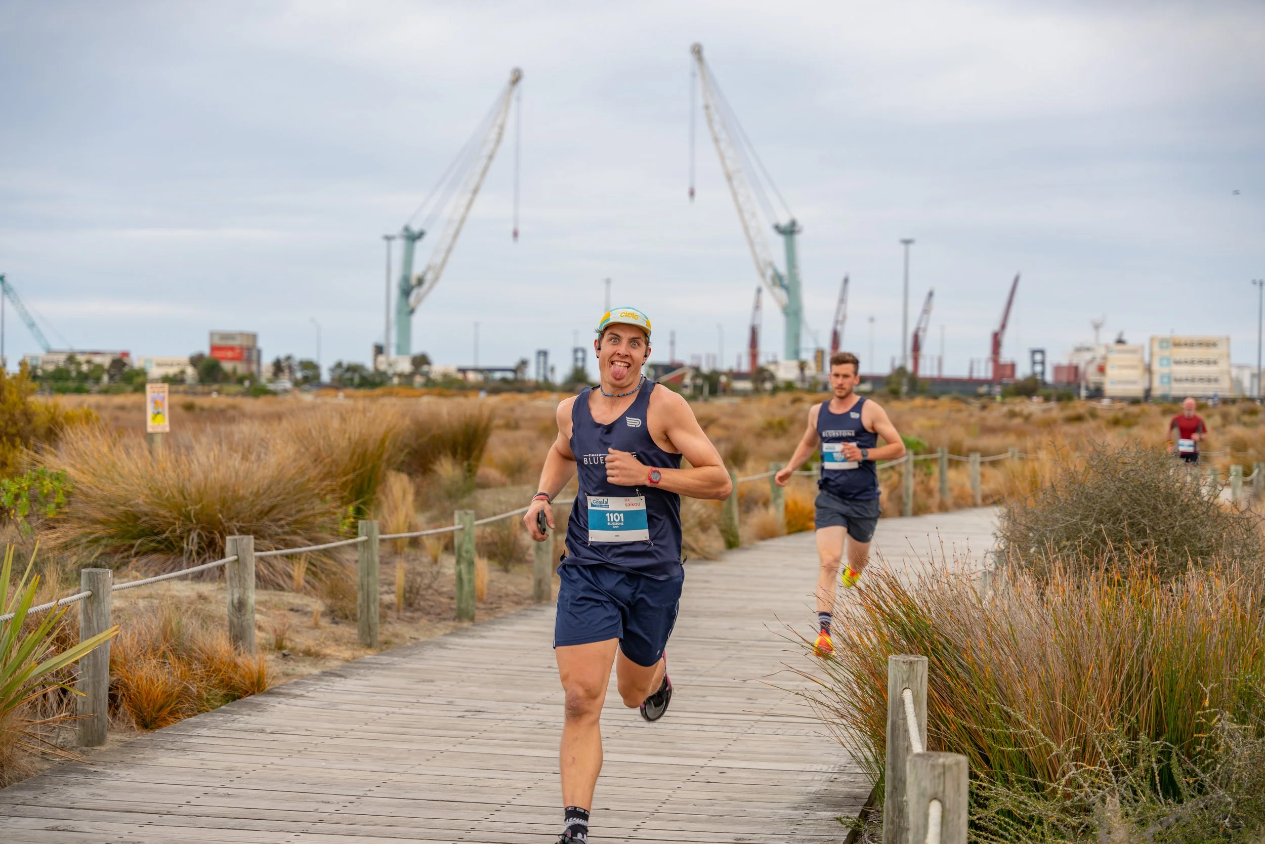 Three runners participating in a race on a wooden pathway in a coastal area with tall grasses, industrial cranes, and containers in the background. The lead runner is sticking out his tongue and wearing a blue tank top, shorts, a cap, and a race bib 