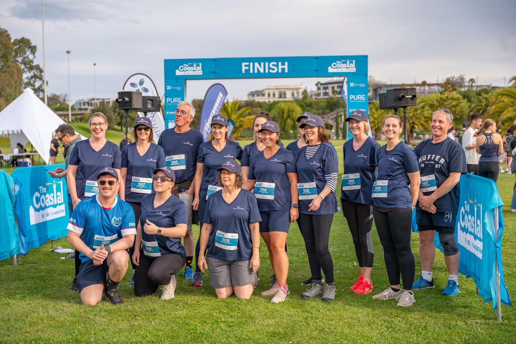 Group of runners in navy and blue shirts posing at the finish line of a marathon at The Coasal event.