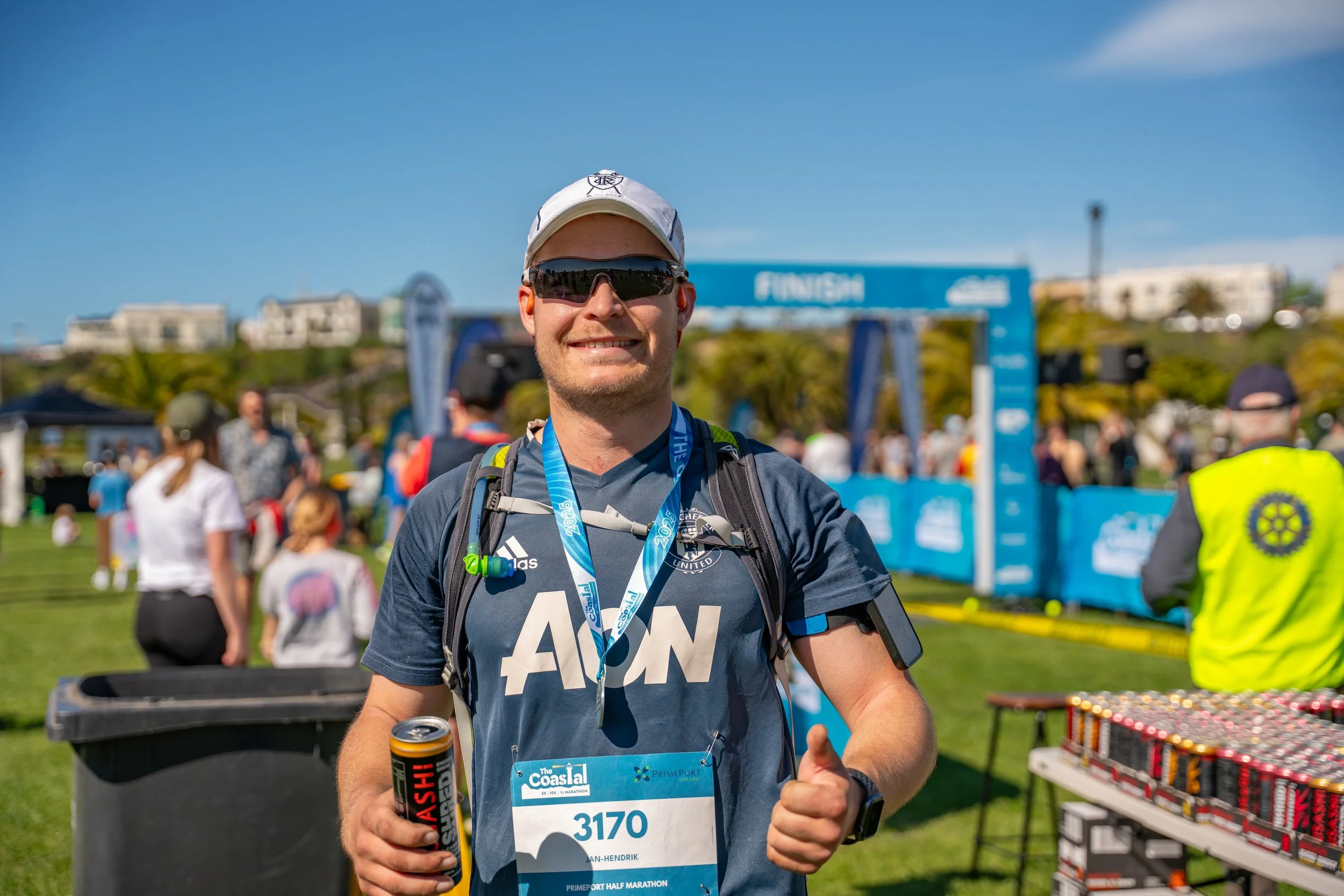 Man with a medal at a marathon race finish line, holding a can, smiling, wearing sunglasses and sports gear, in a sunny outdoor park setting.