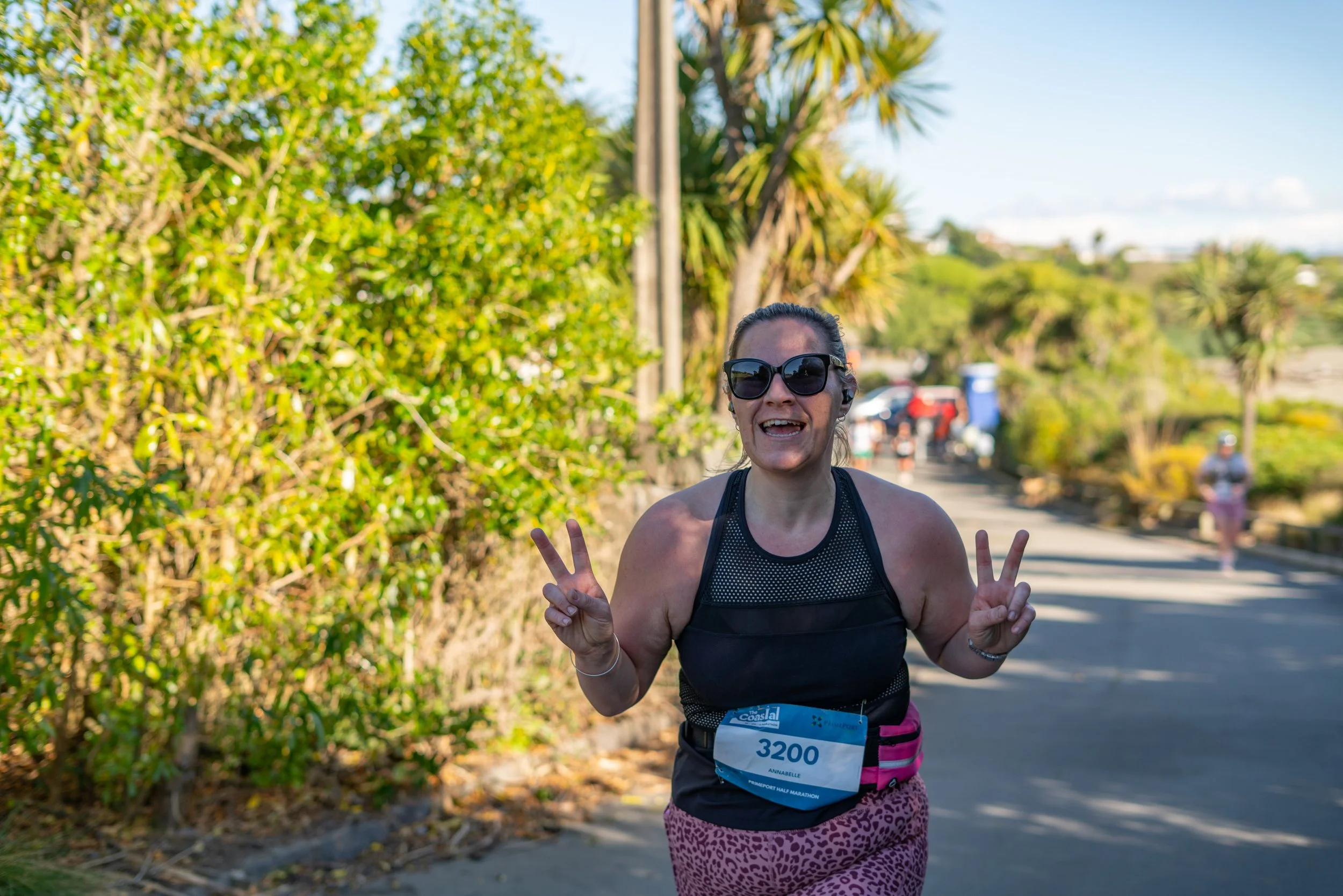 Woman smiling and showing peace signs while running in a race, wearing sunglasses and a black athletic top, with a race bib numbered 3200.