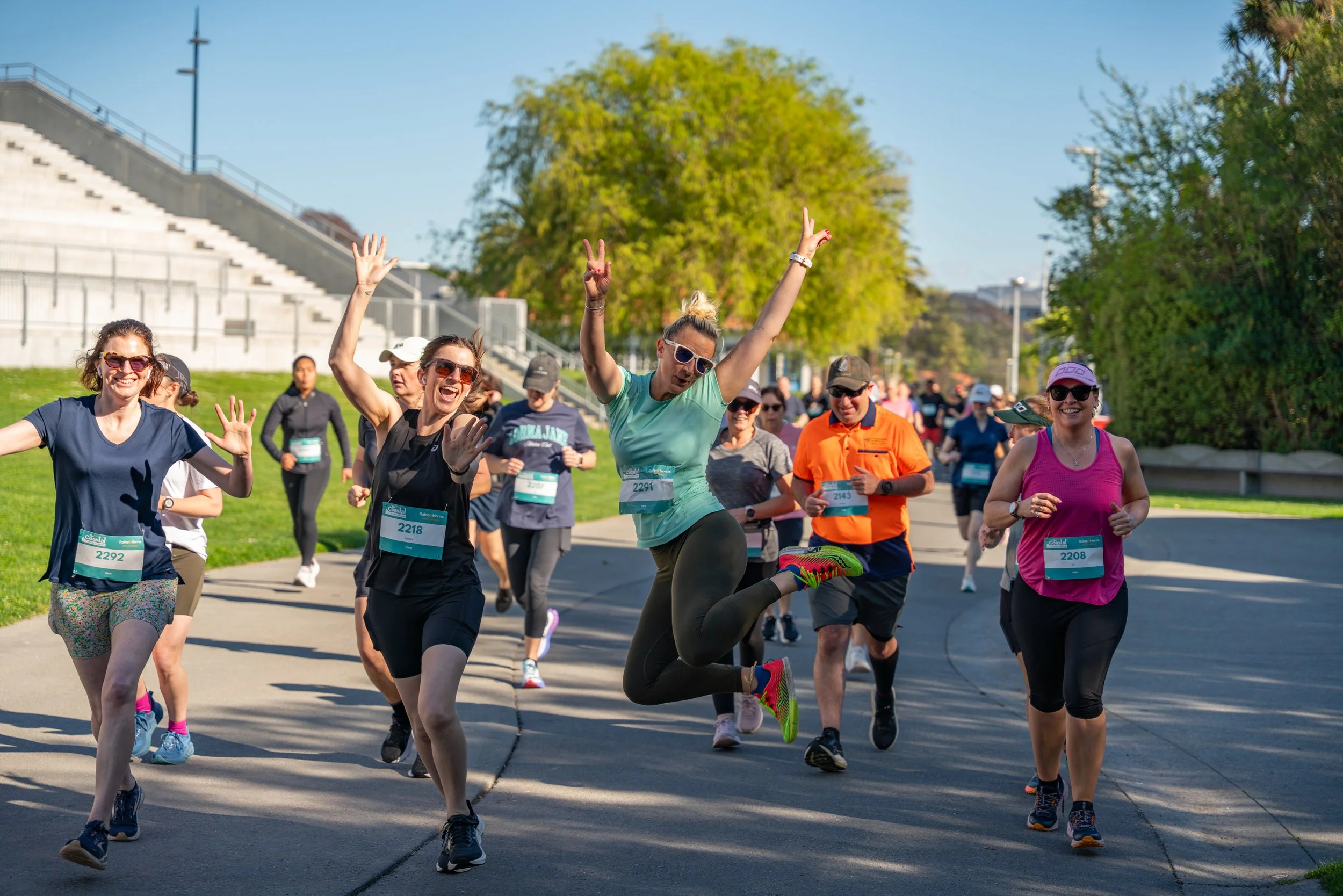 Group of marathon runners smiling and running on a sunny day with trees and a stadium in the background.