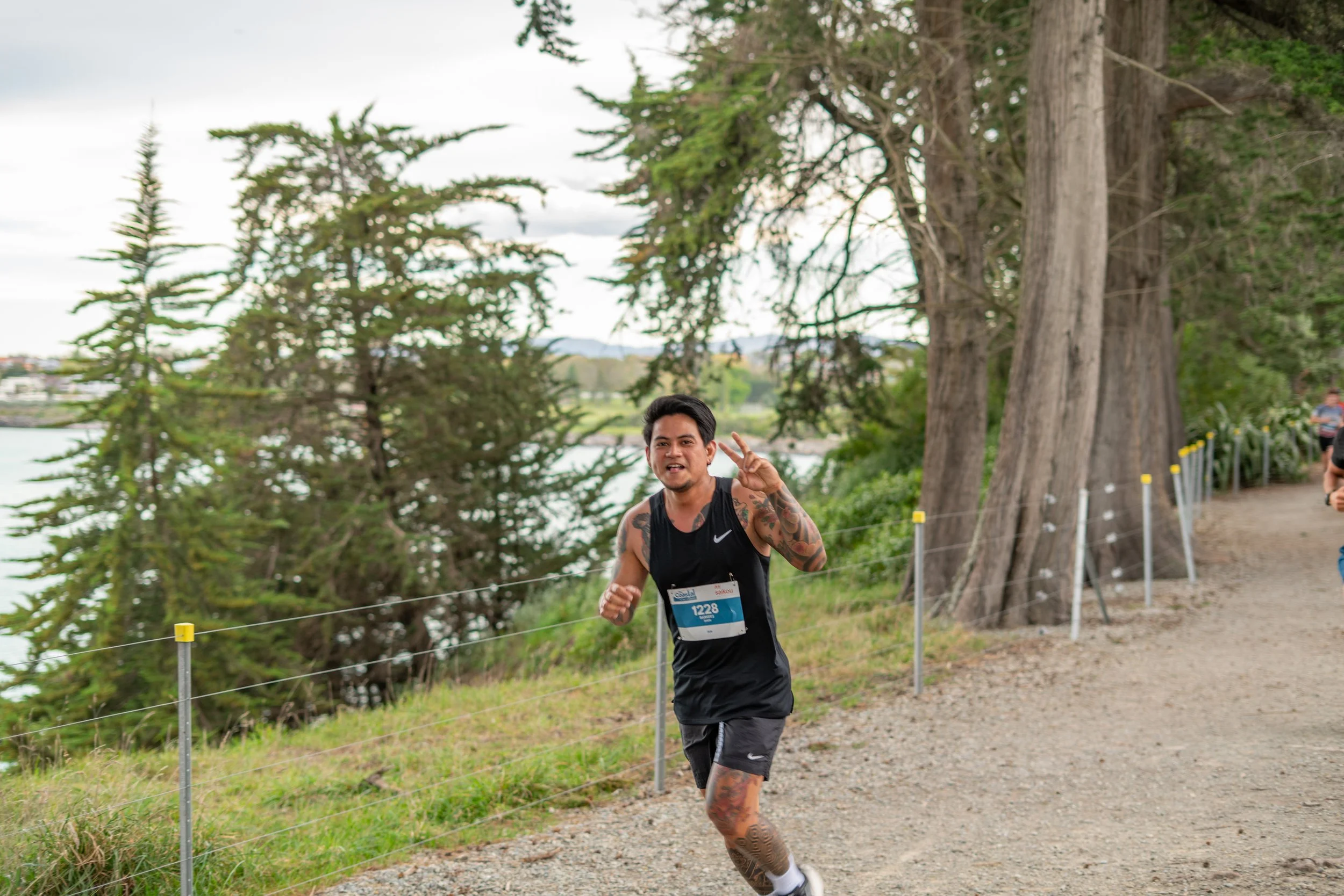 A man running on a trail near a lake with trees in the background, making a peace sign and smiling.