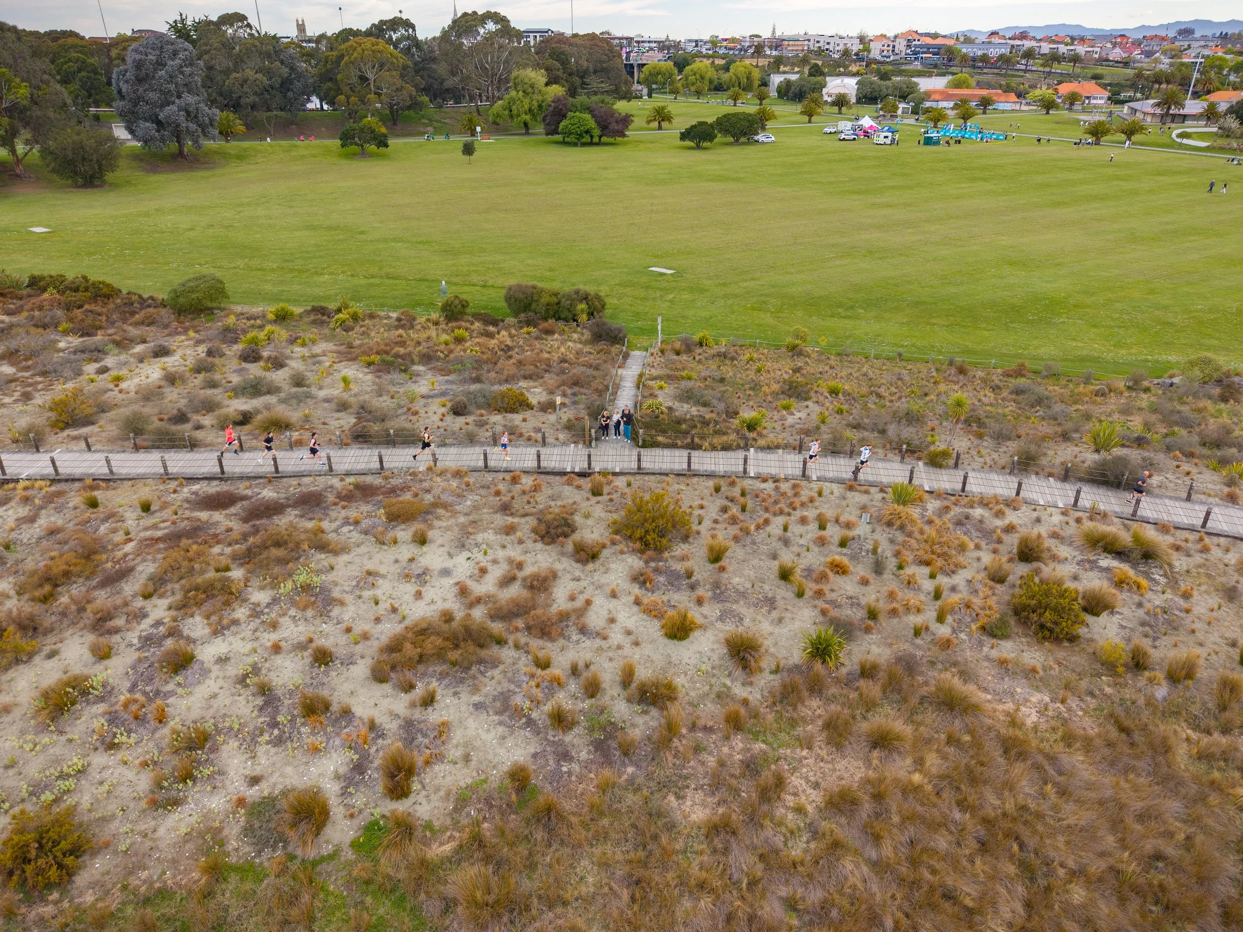 An aerial view of a coastal park with sandy, desert-like terrain and sparse vegetation in the foreground, a paved walkway with people walking, and lush green grass, trees, and buildings in the background.