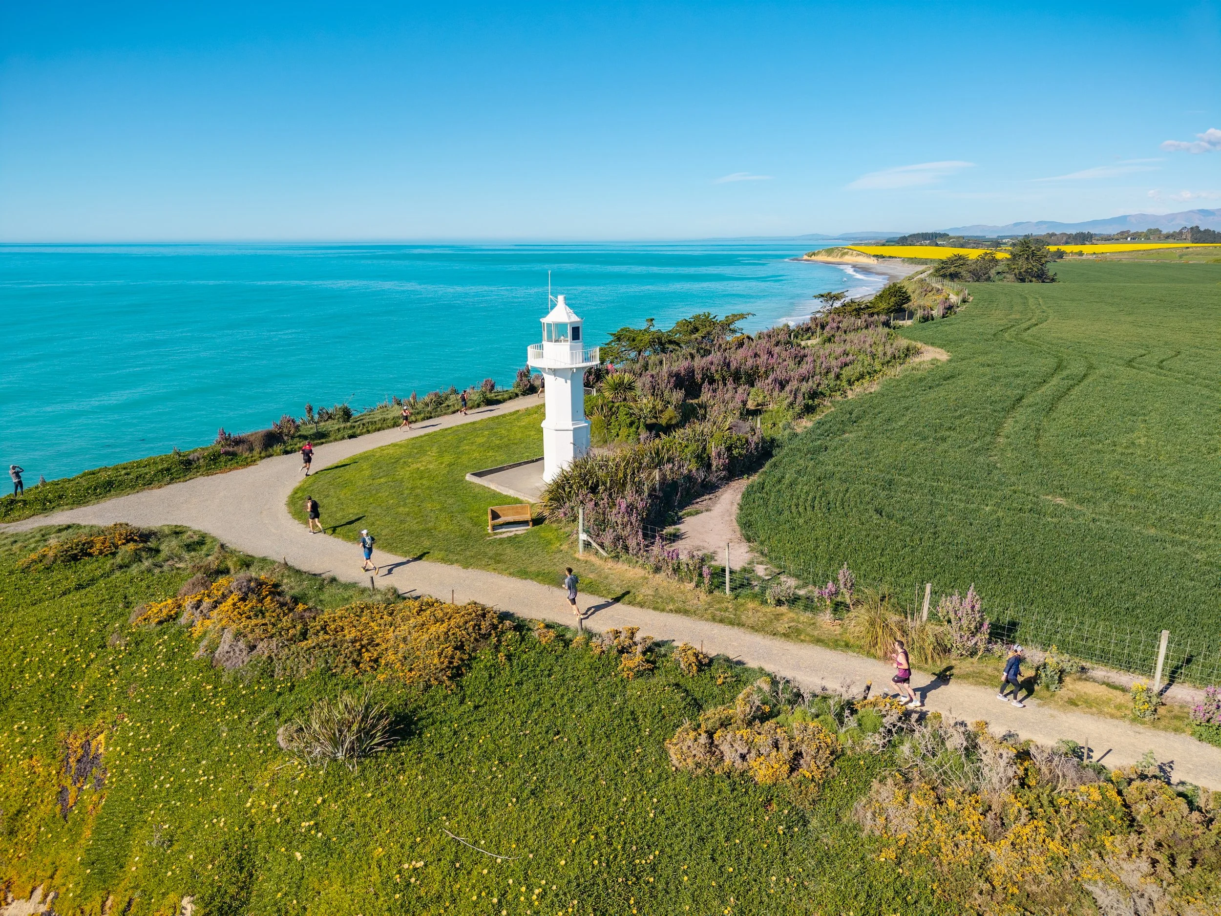 Aerial view of a coastal park with a white lighthouse, walking trail, green fields, and a blue ocean under a clear sky.