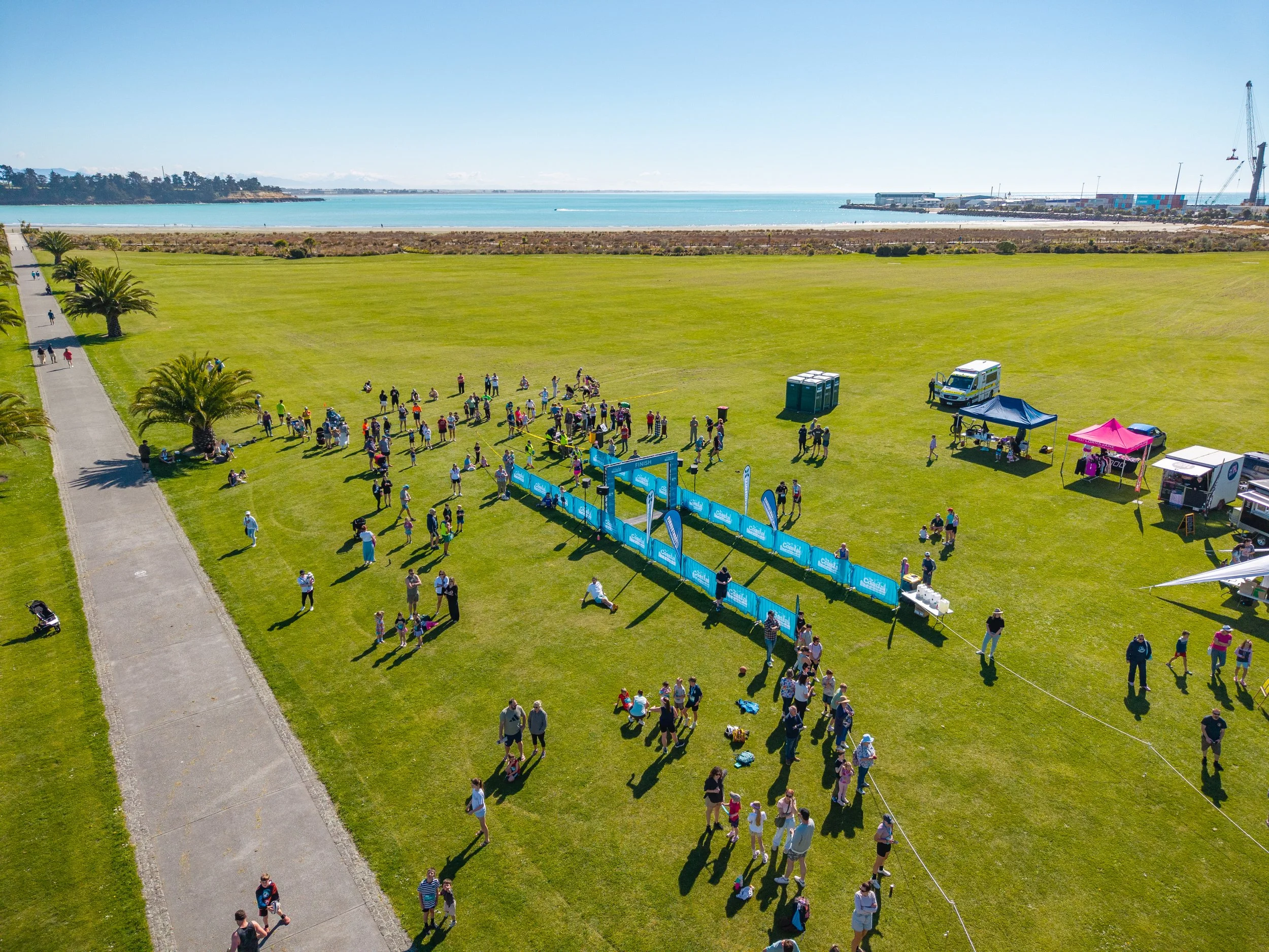 Aerial view of an outdoor event on a grassy field near the ocean, with people walking and gathered around the finish line, tents, and vendor booths.