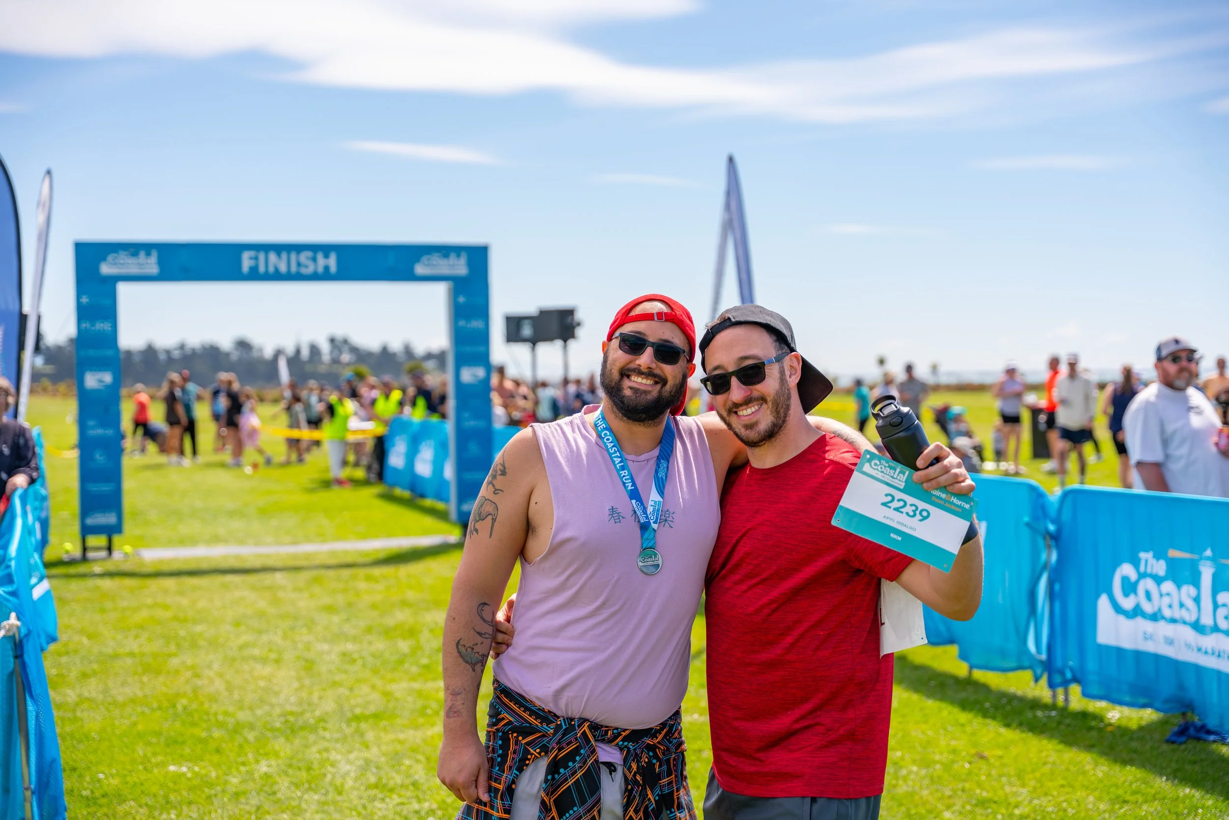Two smiling men celebrating at the finish line of a race, one holding a water bottle and race bib number 2239, surrounded by other participants and spectators outdoors on a sunny day.
