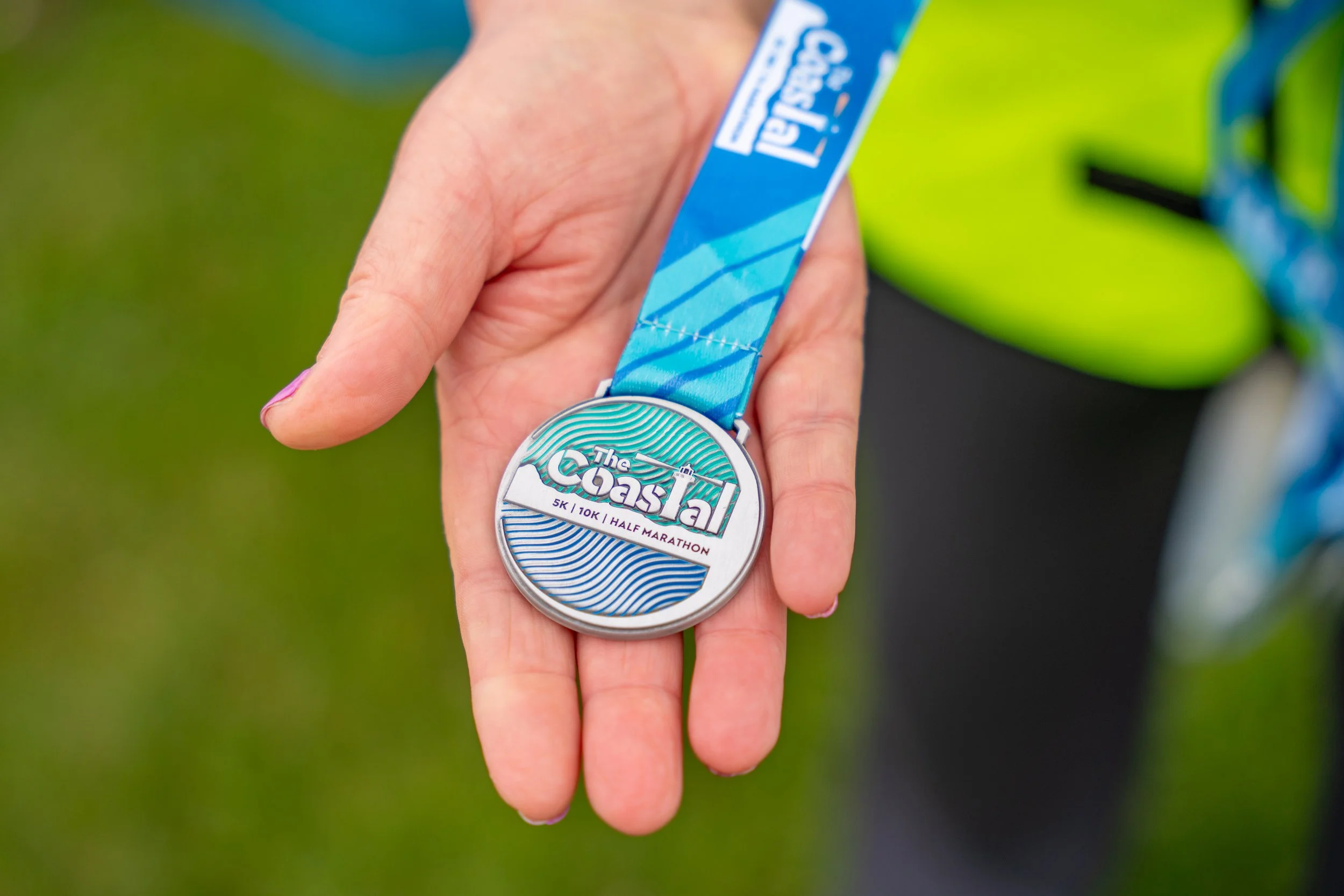 Close-up of a hand holding a medal for The Coastal 5K, 10K, Half Marathon with a blue and green ribbon, against a grassy background.