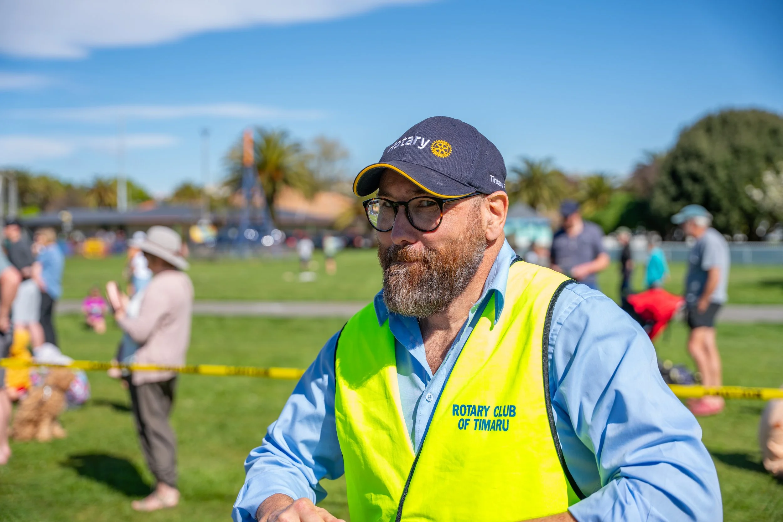 A man wearing a blue cap with Rotary logo, glasses, and a yellow vest with 'ROTARY CLUB OF TIMARU' text, standing outdoors at a community event on sunny day with other attendees and trees in background.