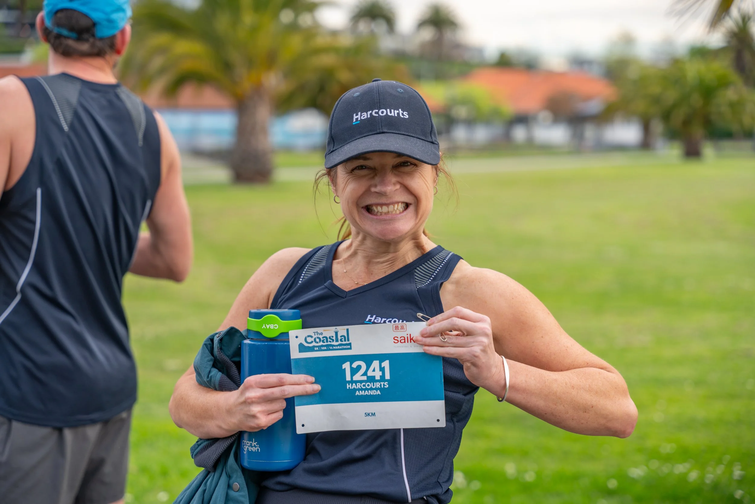 A woman at a race event smiling and holding a race bib and small items, with another runner nearby, in a park with green grass and trees.