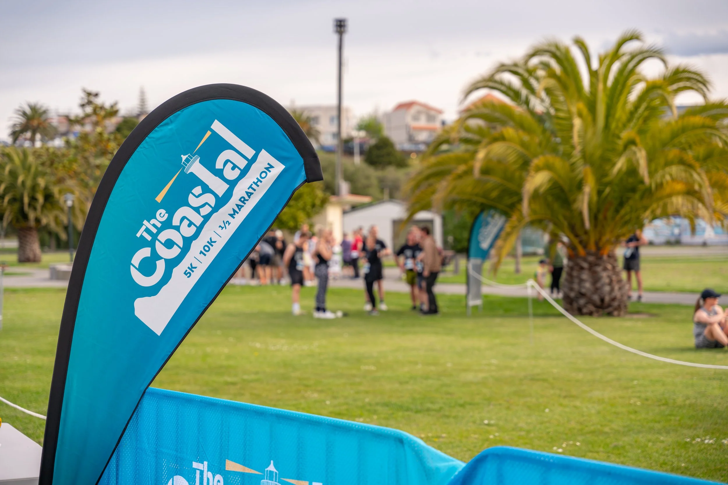 A blue flag with the text "The Coast" and logos for a marathon, half marathon, 10K, 5K, and 1K, on a grassy area with a group of people gathered in the background, palm trees, and cloudy sky.