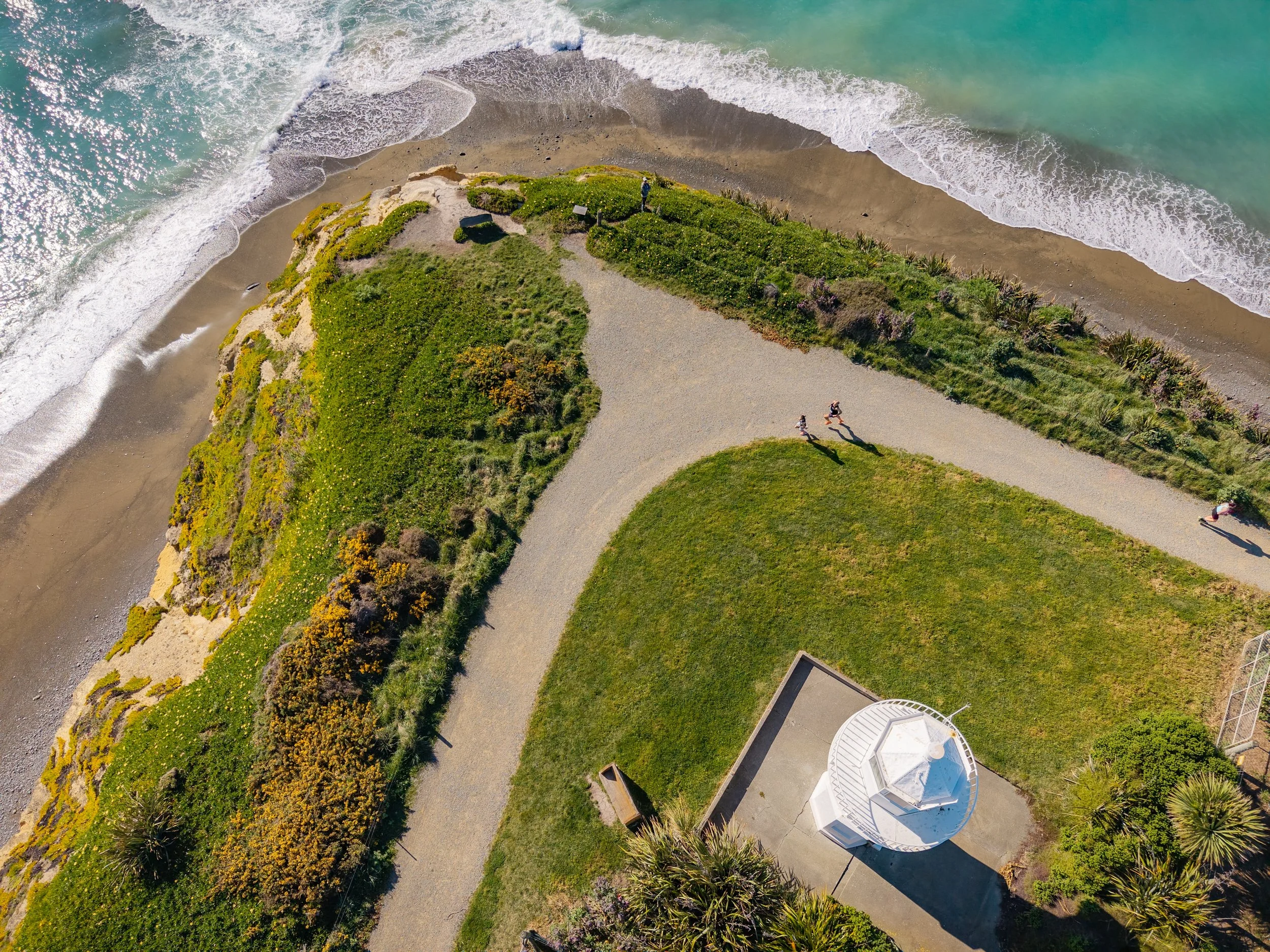 An aerial view of a coastline with a pebble pathway, green grass, and a small white lighthouse. The beach has waves crashing onto the sand, and several people are walking along the path.