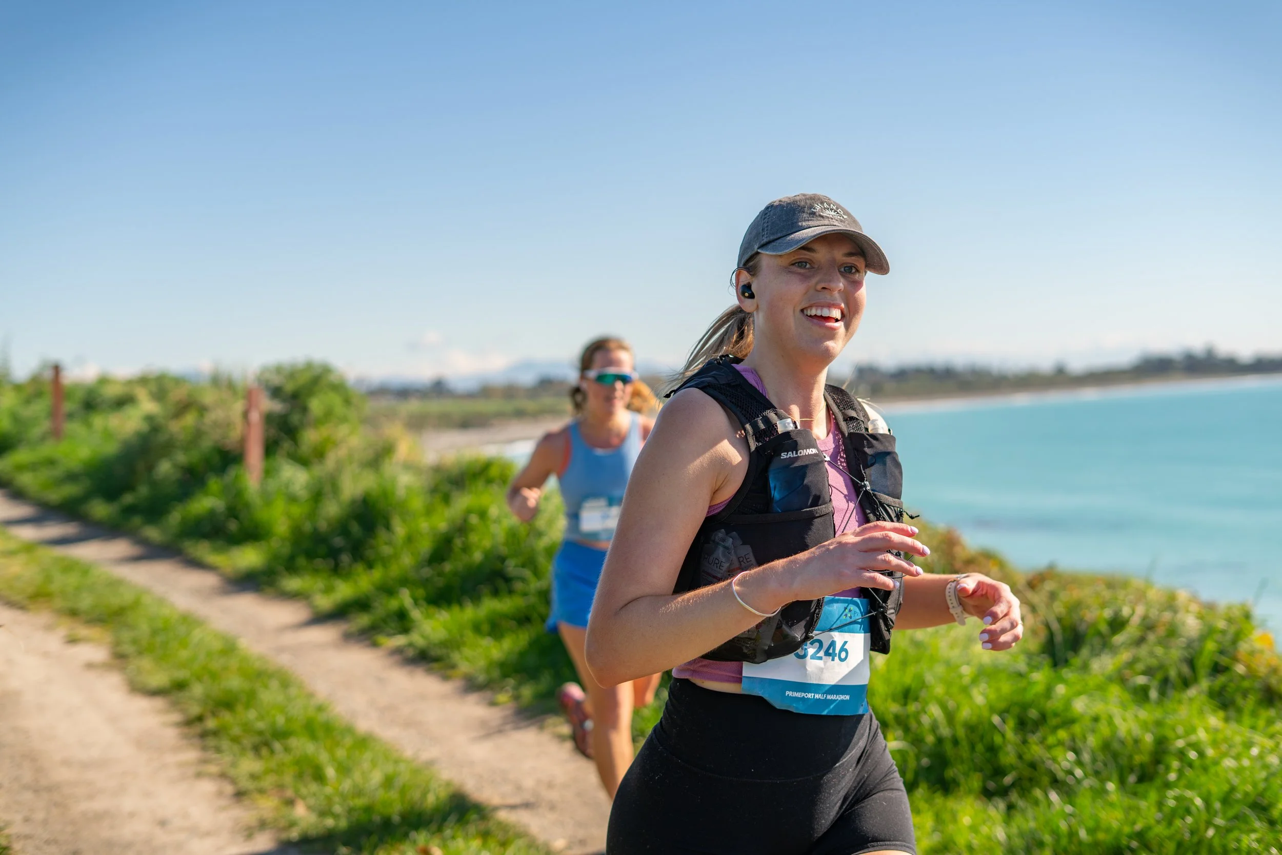 Two women running along a coastal trail with water and greenery in the background on a sunny day.