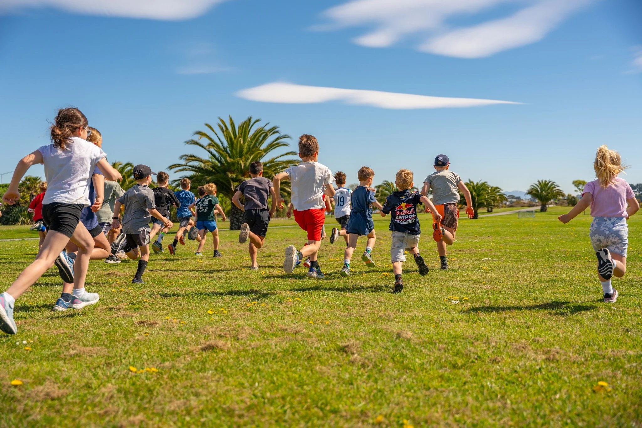 Children running on a grassy field under a blue sky with palm trees in the background.