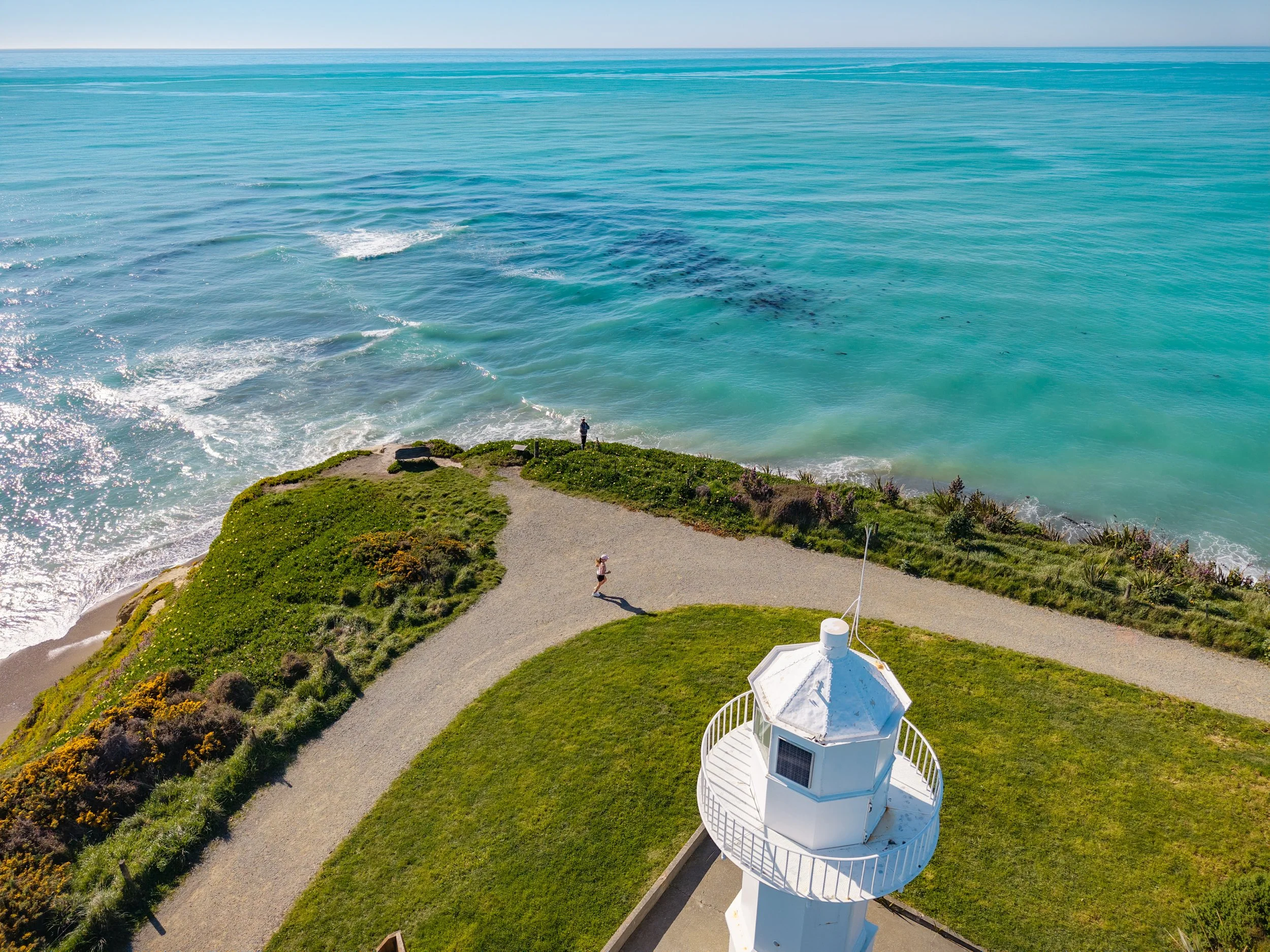 An aerial view of a lighthouse on a grassy area near the coast with a person jogging along a winding path, while another person stands near the water's edge, with the ocean stretching to the horizon.