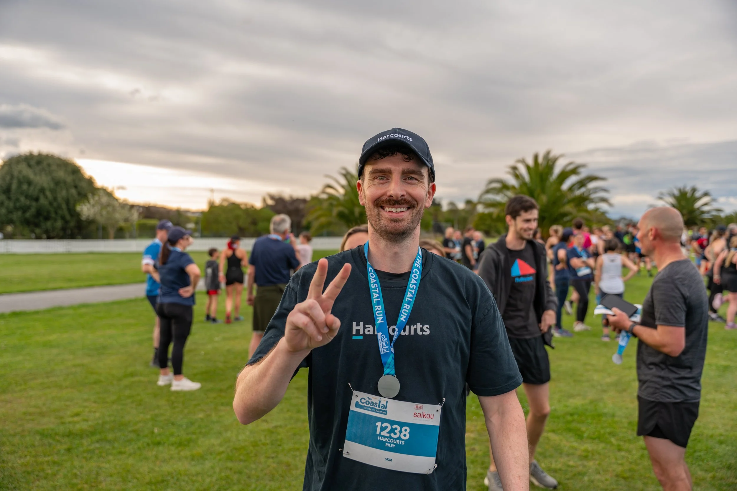 A smiling man wearing a black shirt, cap, and medal around his neck posing for a photo at a race event. He is holding up a peace sign with his right hand, surrounded by other people at the race afterparty on a grassy field with palm trees and cloudy 
