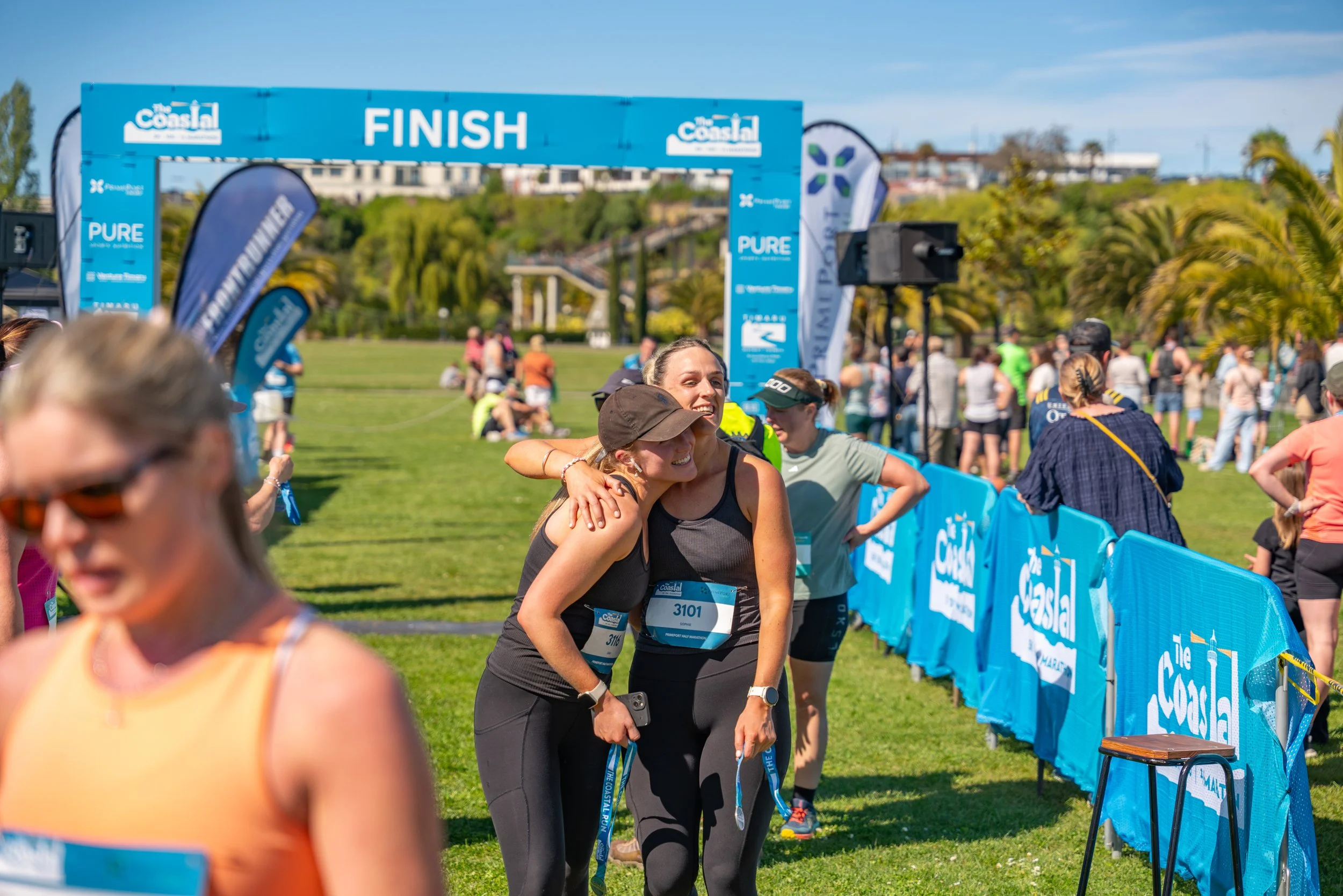 Two women in athletic clothing hugging at the finish line of a race, with a blue finish line banner overhead and a crowd of people in the background.
