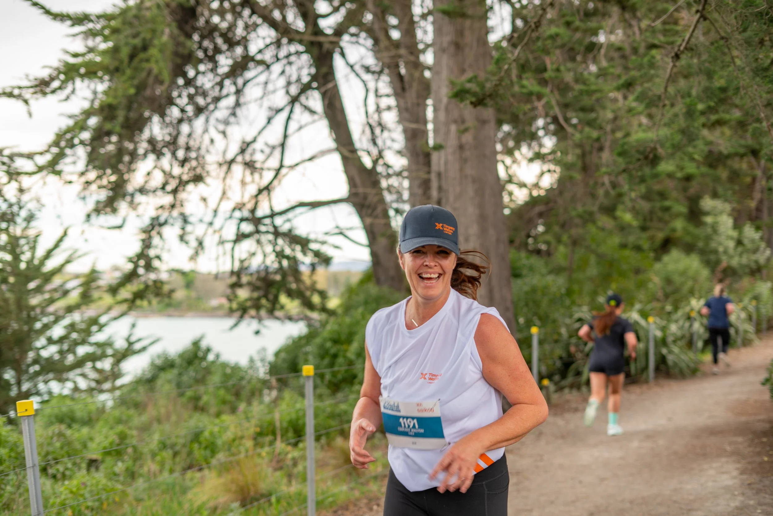A smiling woman running outdoors in a race, wearing a white sleeveless shirt, black shorts, and a dark cap, with trees and other runners in the background.