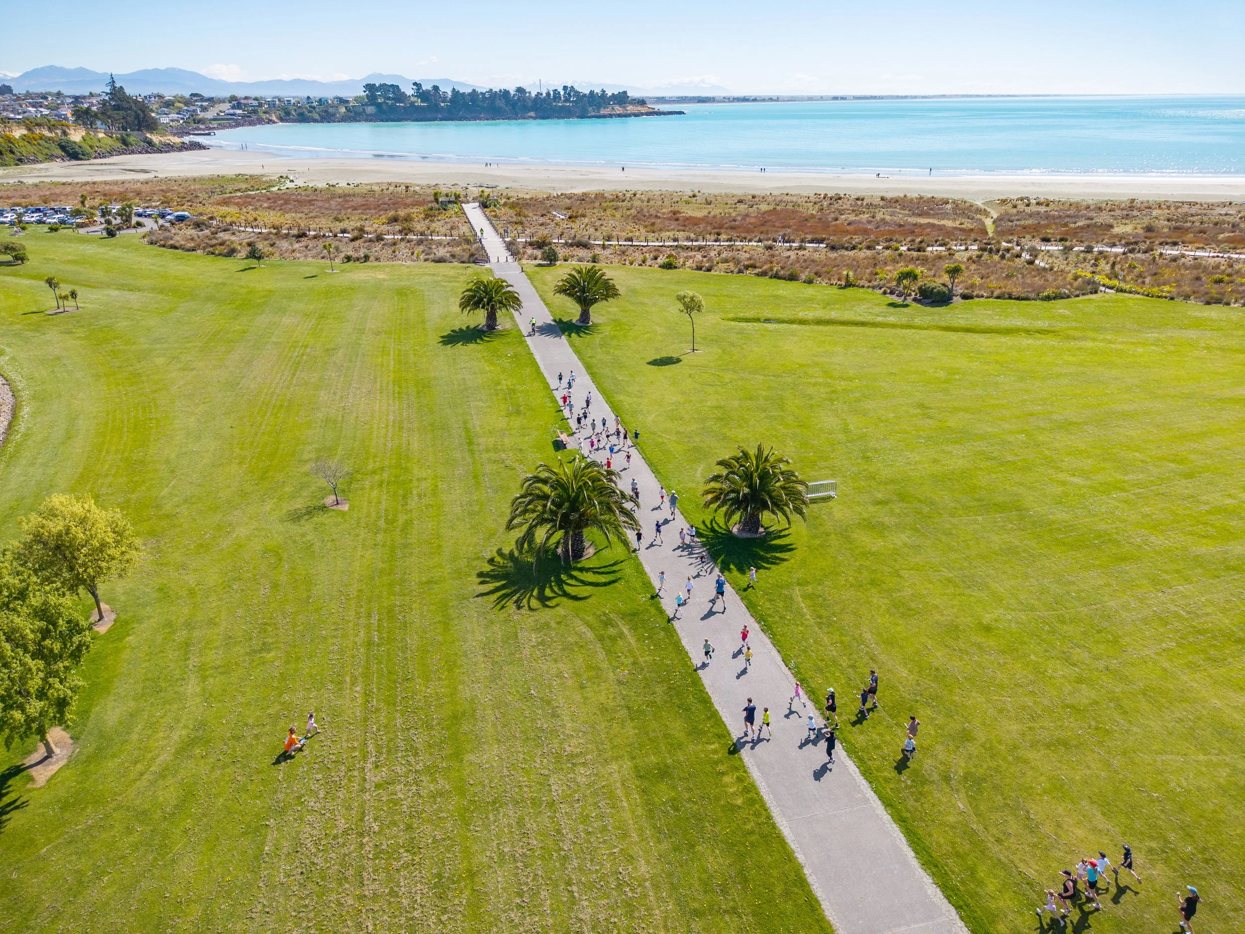 An aerial view of a park near the beach with a paved pathway, palm trees, people walking, and the ocean in the background.