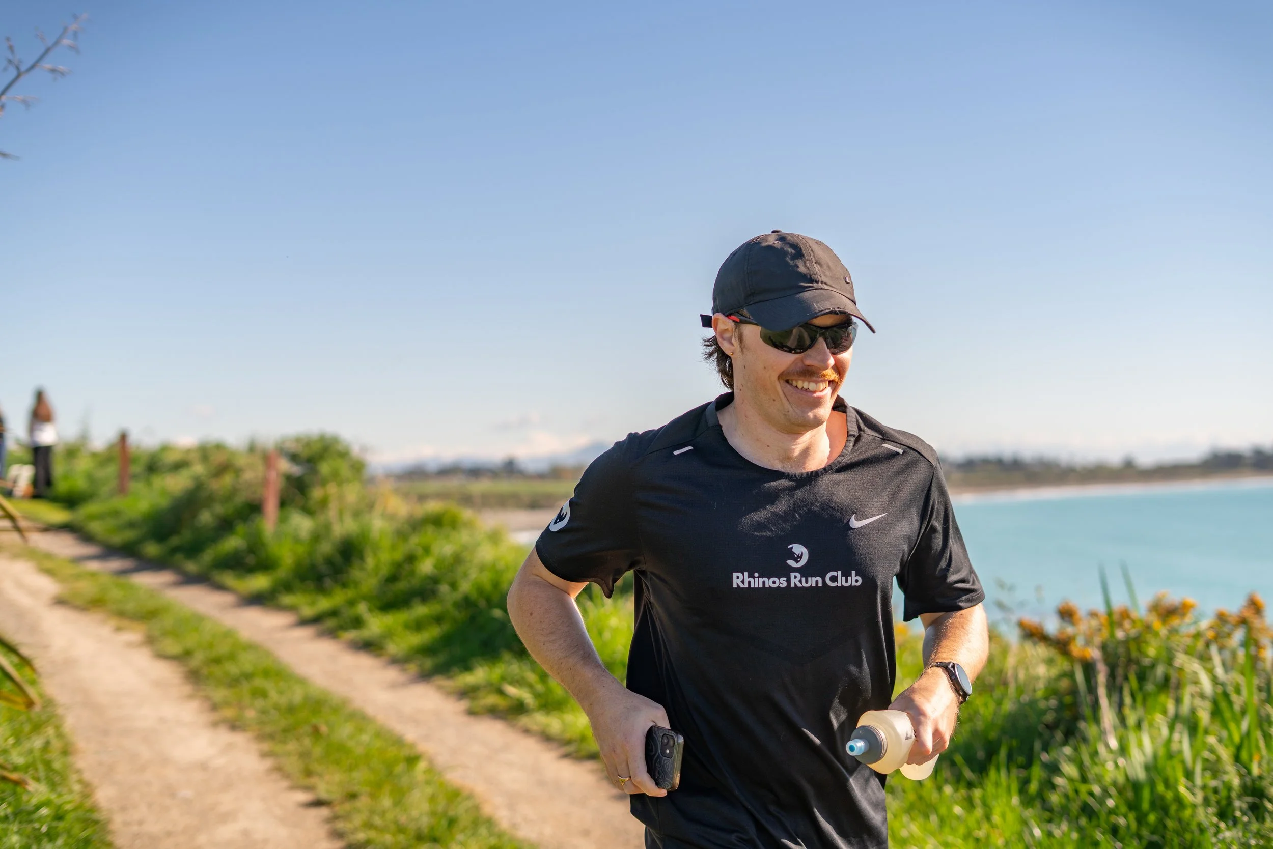 Man running outdoors by water, wearing sunglasses, a cap, and a black Rhinos Run Club shirt, smiling, holding a water bottle and smartphone.